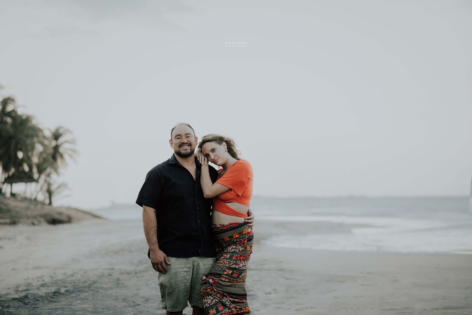 A couple embraces warmly on a tropical beach with palm trees and ocean waves in the background.