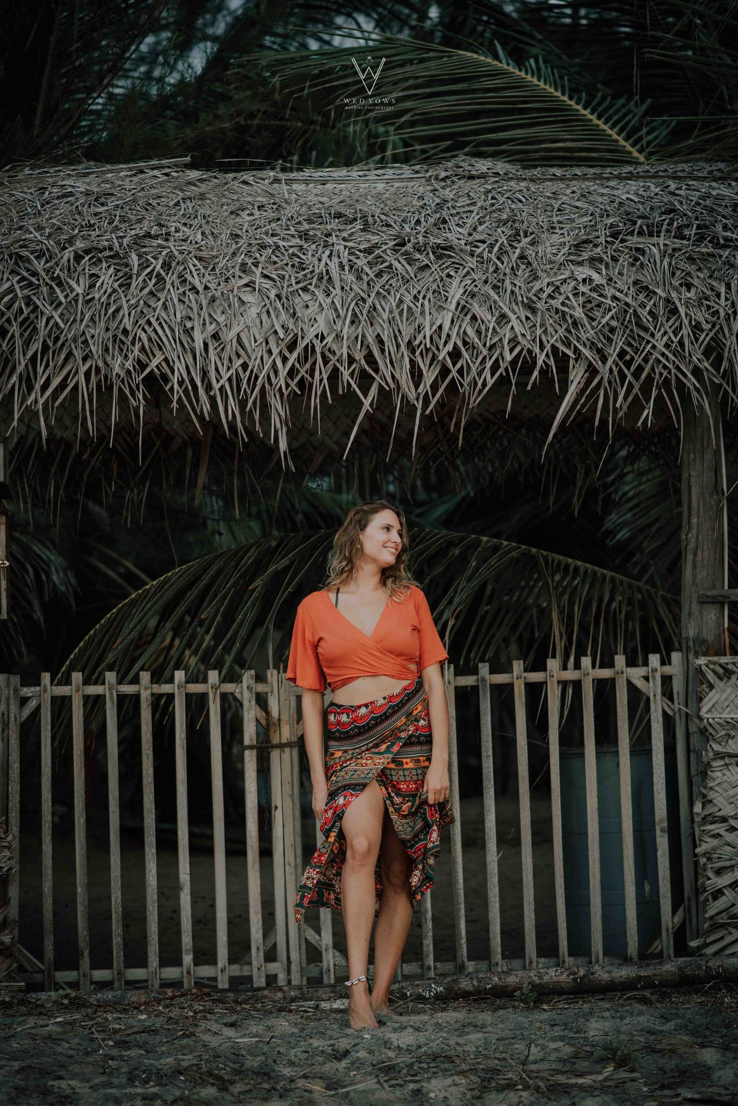Woman in orange wrap top and colorful skirt posing by a rustic bamboo fence with palm trees.