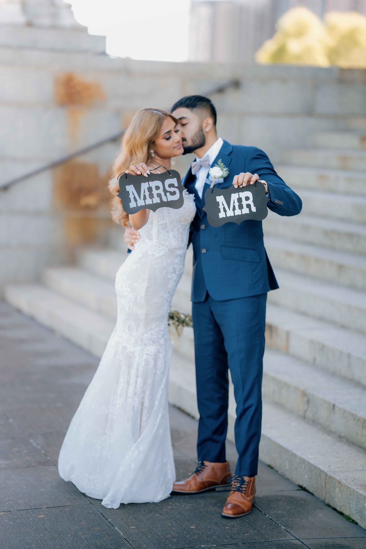 Newlyweds hold MR and MRS signs while sharing a kiss on stone steps.