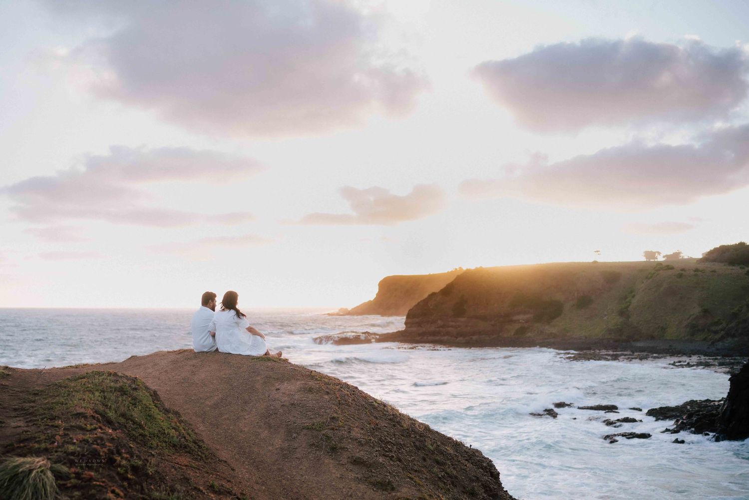 Couple sitting together on coastal cliffs watching a golden sunset over the ocean.