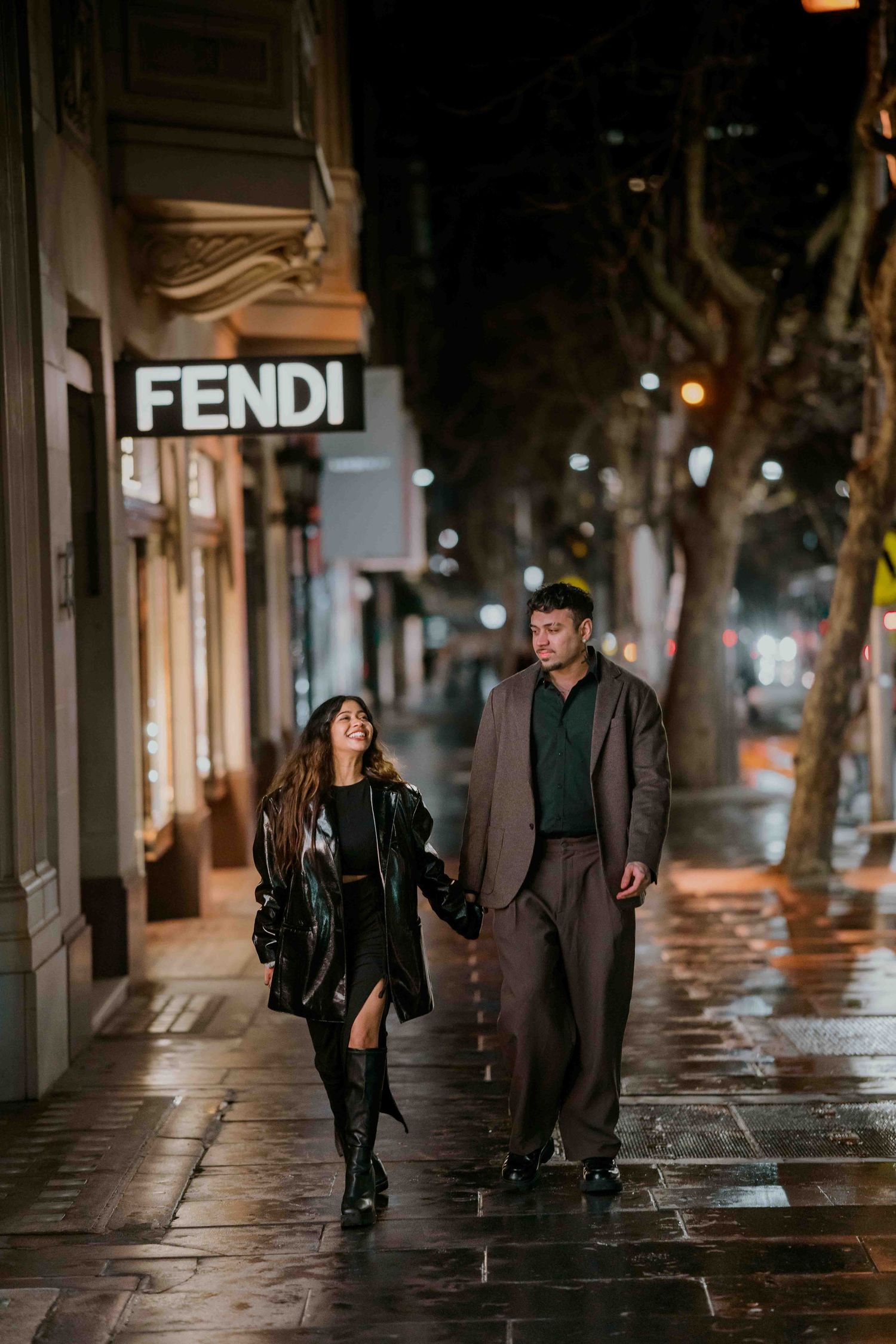 Couple walking hand in hand past a glowing Fendi store sign on a rainy city night.