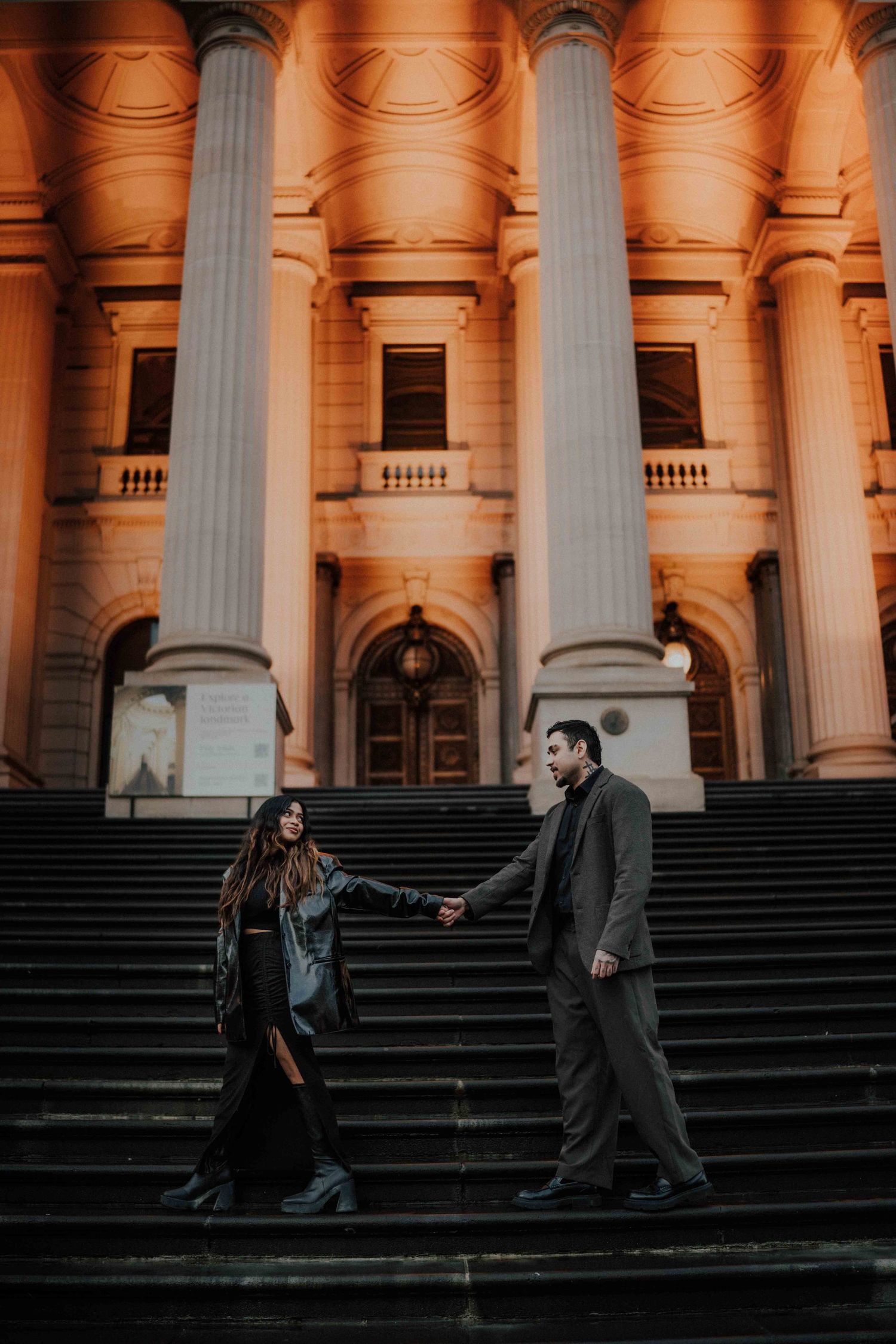 Romantic couple holding hands on steps of grand illuminated classical building with tall columns.
