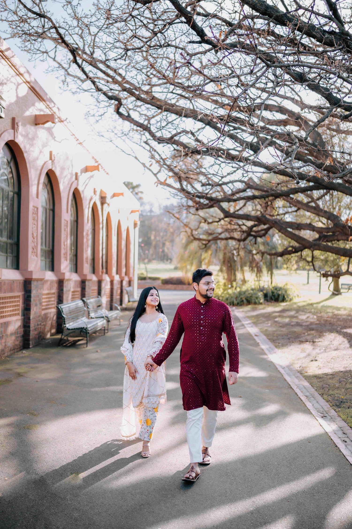 Indian couple in traditional attire holding hands walking along a path beside a red brick building.