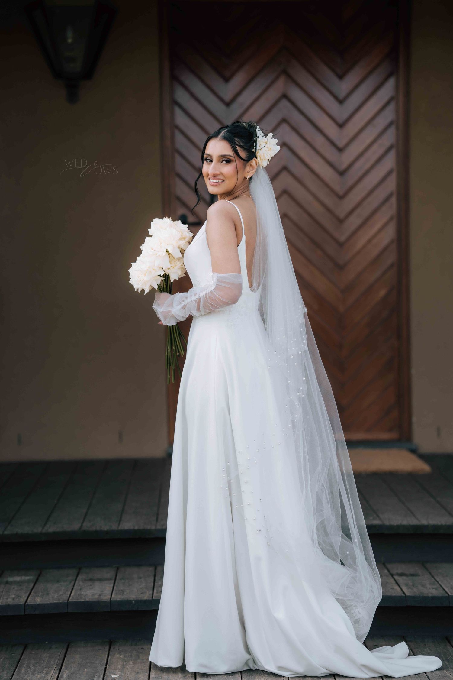 Elegant bride in long-sleeve white gown with veil poses before rustic wooden doors with bouquet.