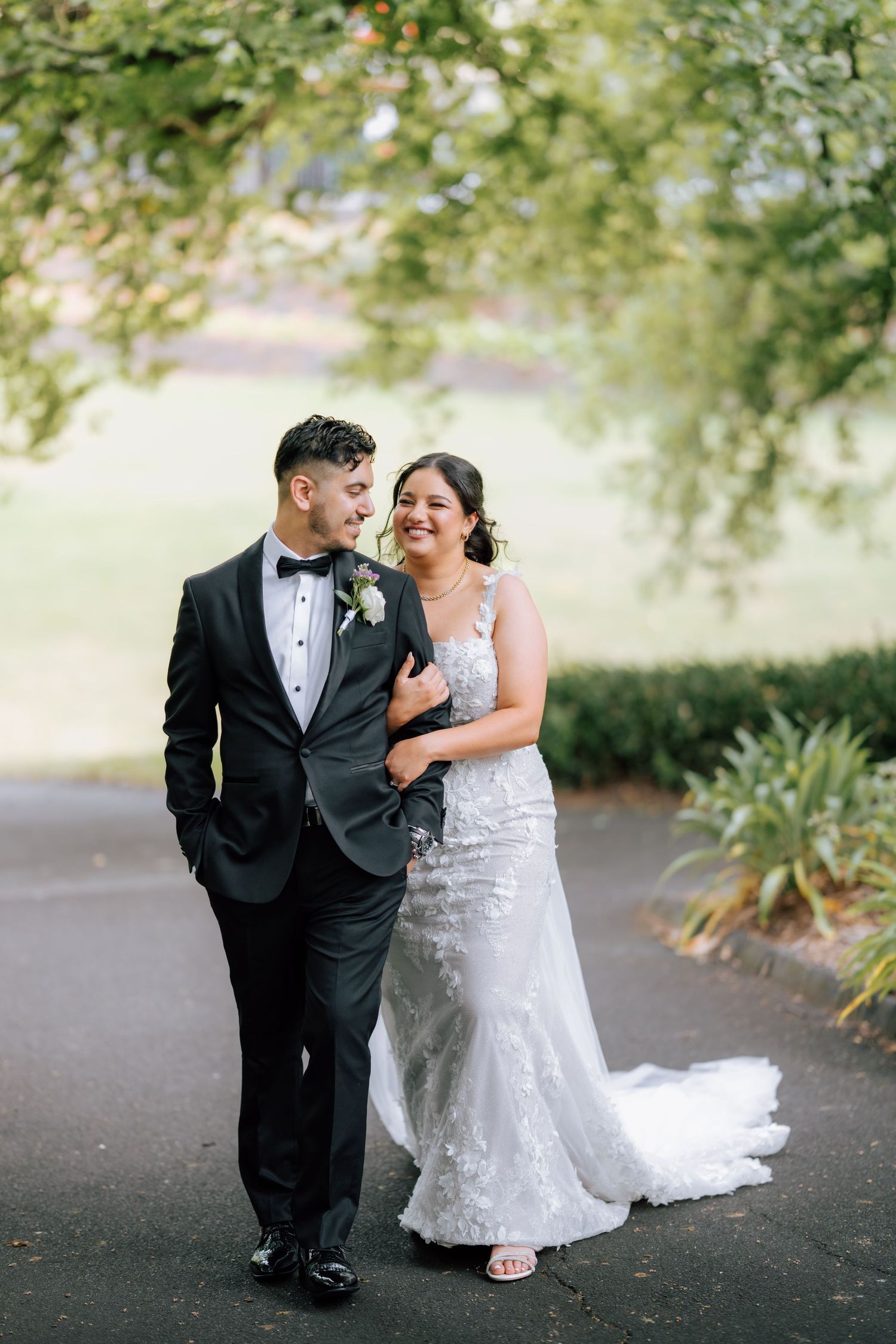 Smiling bride in lace mermaid gown and groom in black tuxedo walk together under tree-lined path.