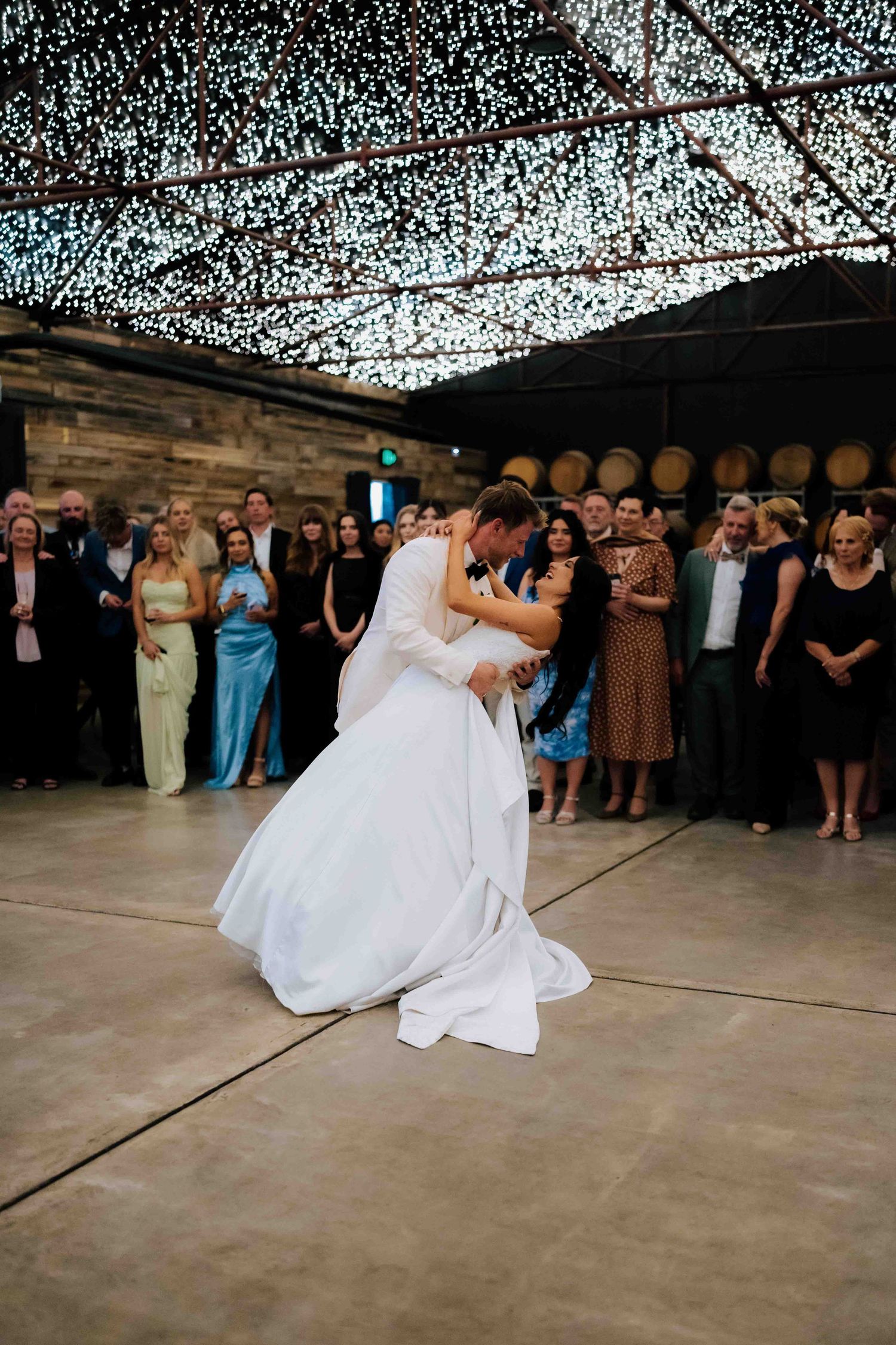 Bride and groom share romantic first dance dip under twinkling fairy lights at winery wedding reception.