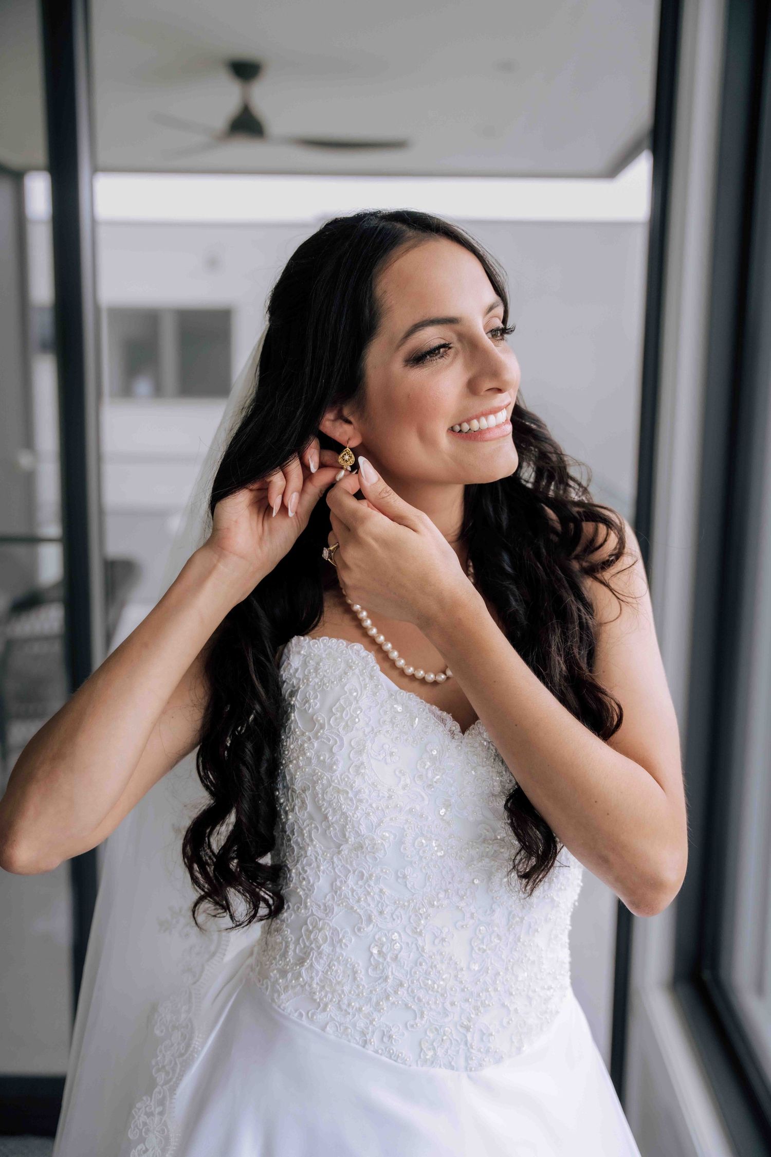 Smiling bride in strapless lace wedding gown adjusts pearl earring while wearing pearl necklace indoors.