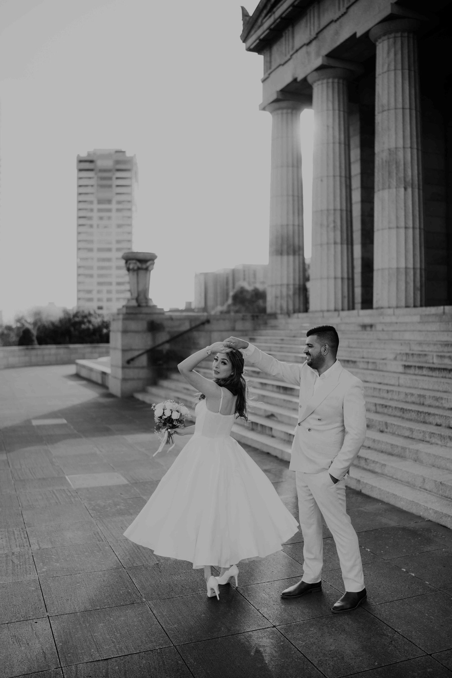 Groom twirls bride in flowing white dress outside grand columned building in black and white wedding photo.