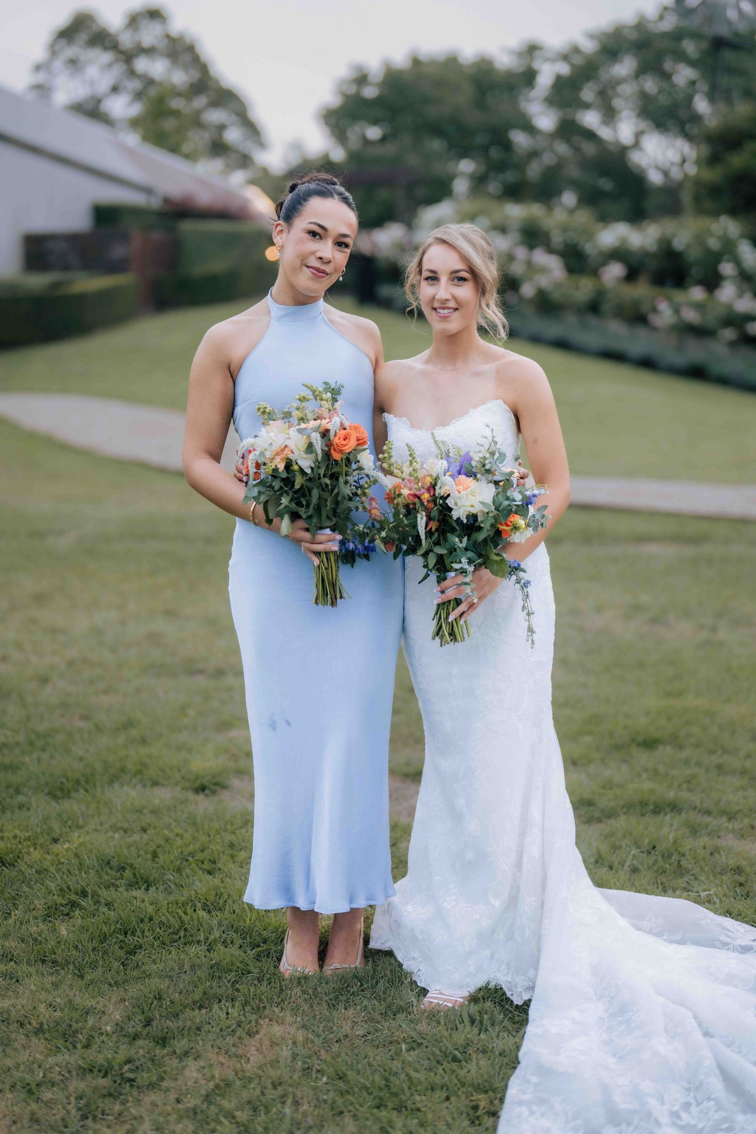 Bridesmaid in light blue halter neck dress poses with bride in white strapless lace gown holding colorful bouquets.