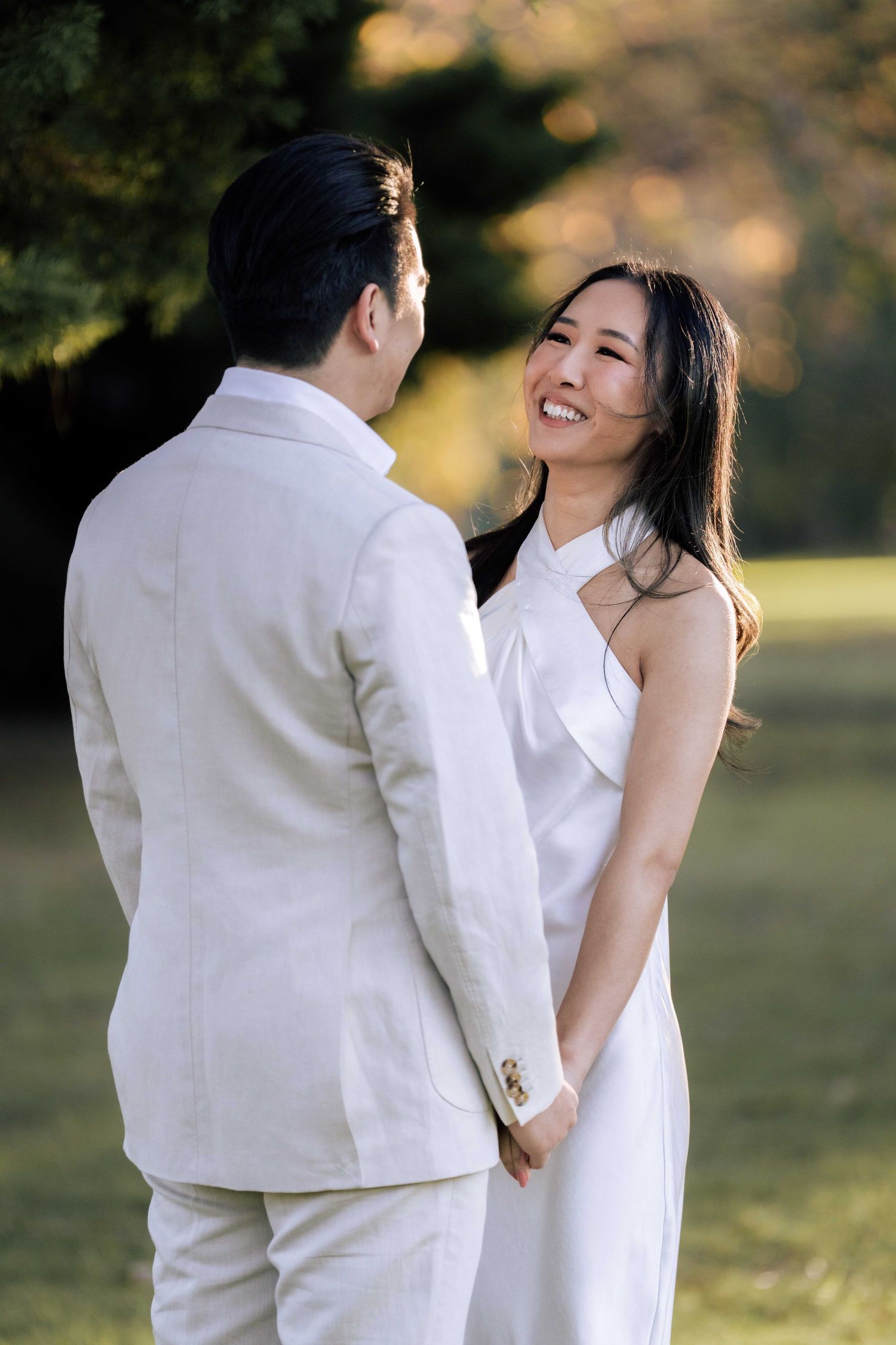 Smiling bride in white halter dress holds hands with groom in cream suit outdoors in a sunlit garden.