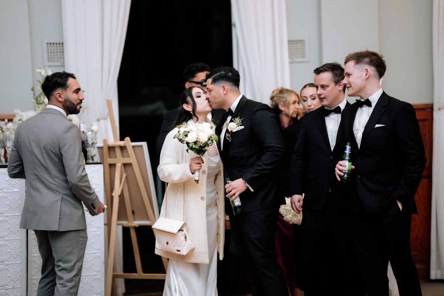 Wedding guests in formal attire mingle and chat near decorative easel at reception.
