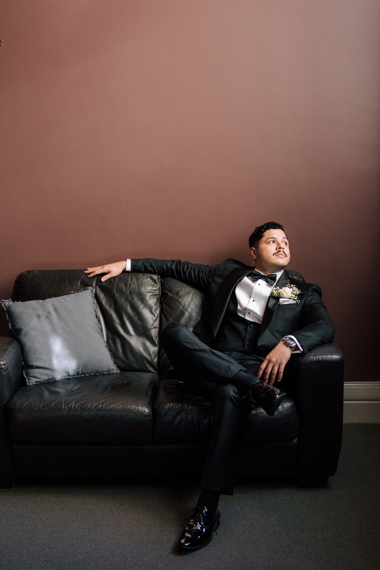 Groom in black tuxedo with boutonniere relaxing on leather sofa before wedding ceremony.