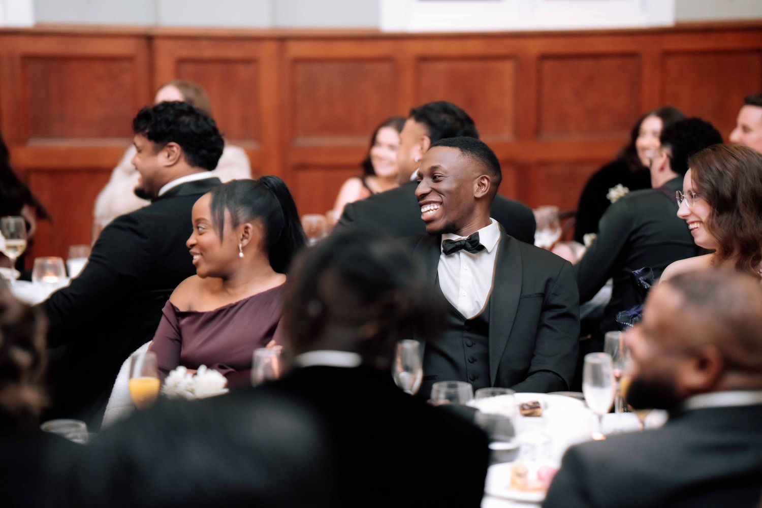 Wedding guests laugh and smile at elegant reception dinner seated at white-clothed tables in formal attire.