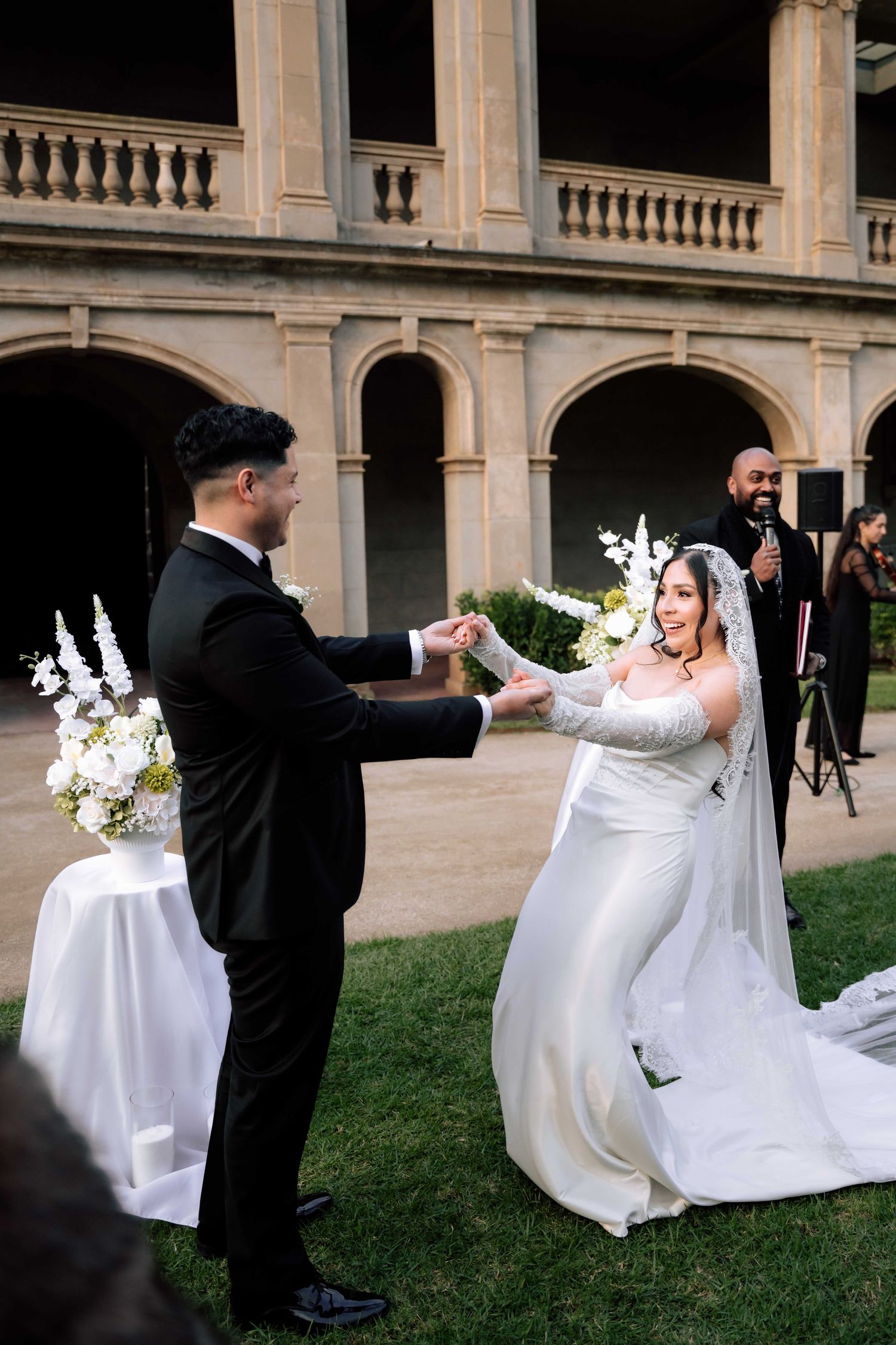 Bride in white gown and groom in black tuxedo hold hands during outdoor wedding ceremony at elegant venue.