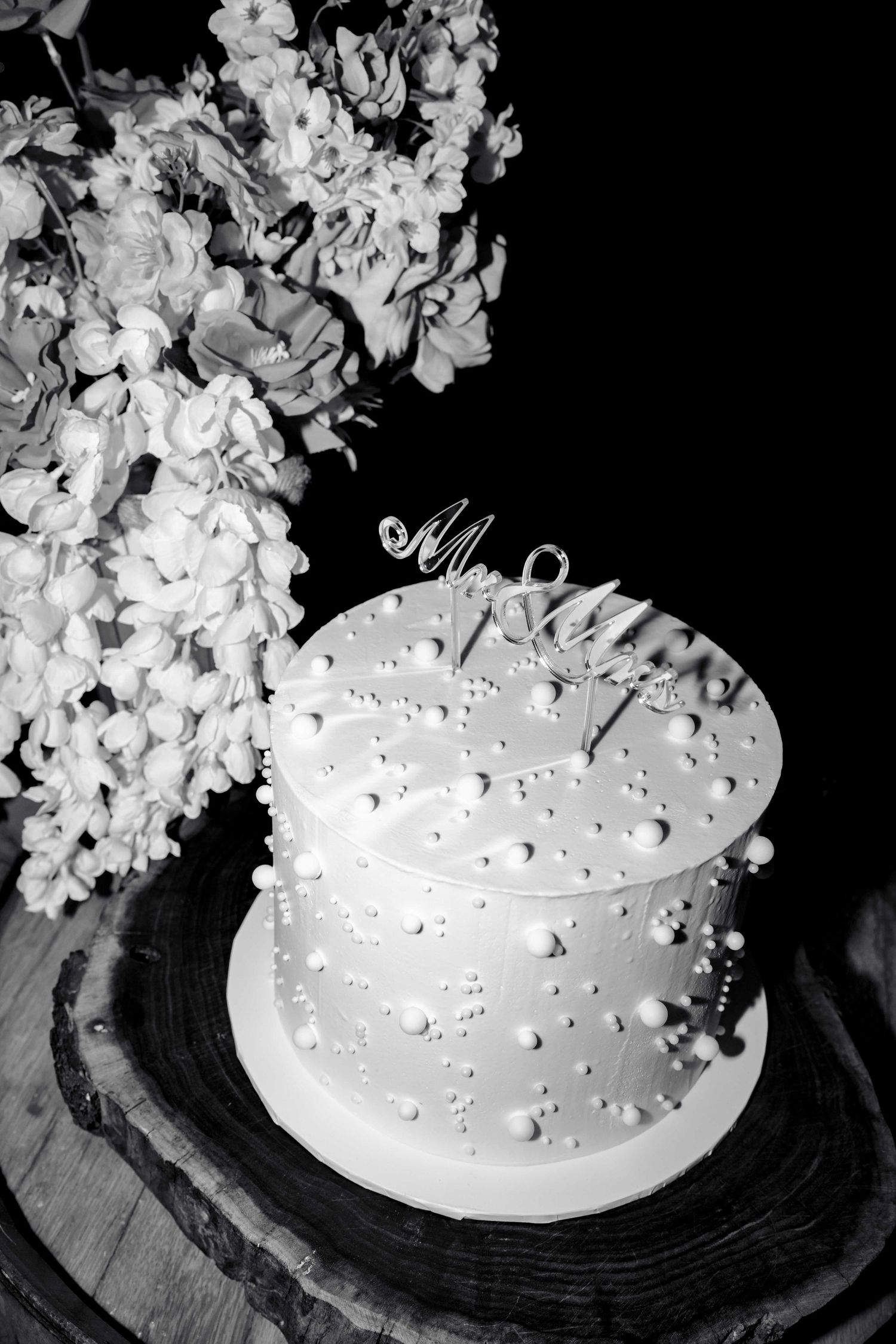 Elegant white wedding cake with pearl details and Mr & Mrs topper beside white floral arrangement, black and white.