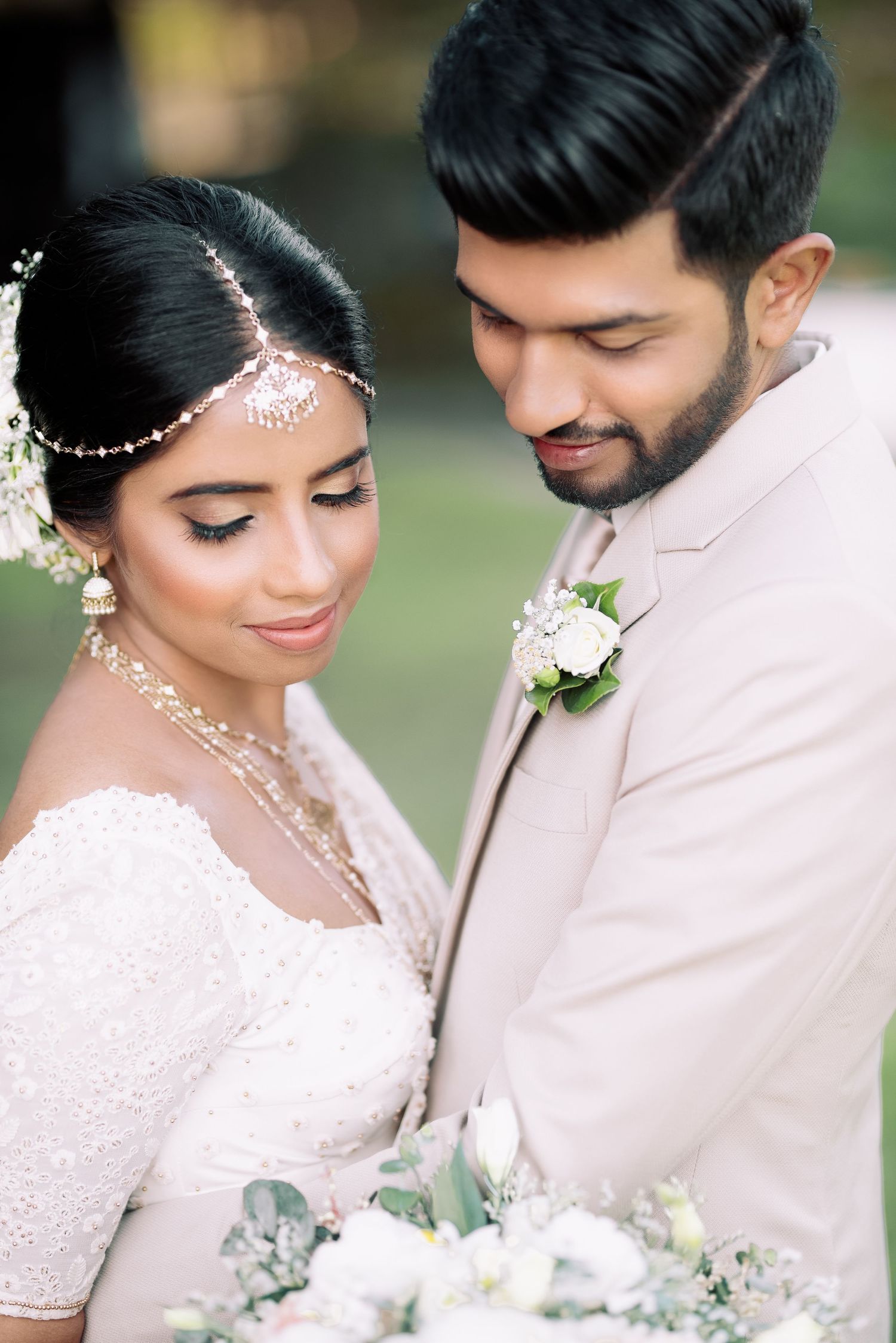Indian bride in lace gown with maang tikka and groom in beige suit with white boutonniere.
