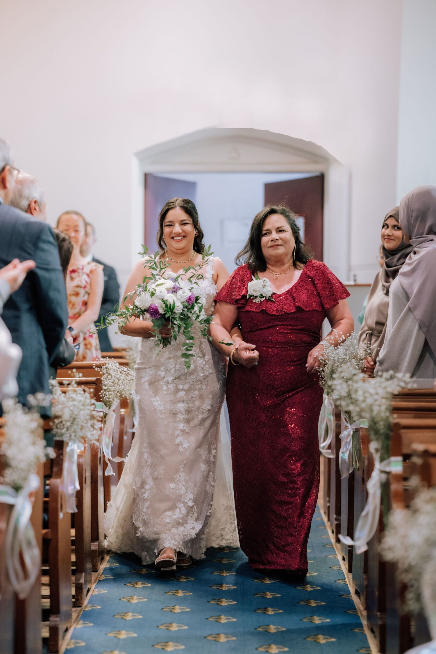 Bride walks down church aisle with mother in red dress holding floral bouquet.