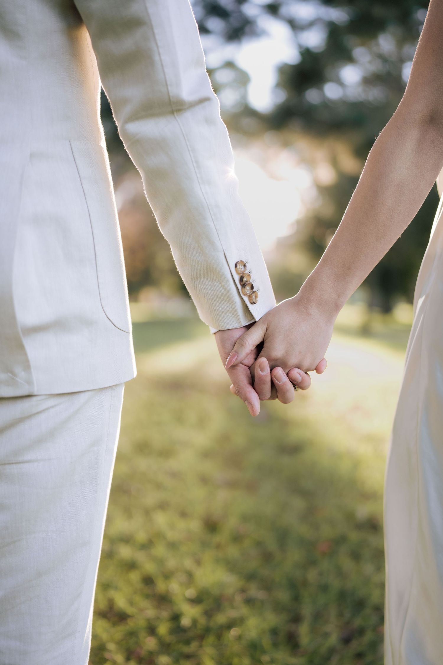 Close-up of a wedding couple holding hands in a lush green park, both dressed in elegant white outfits.