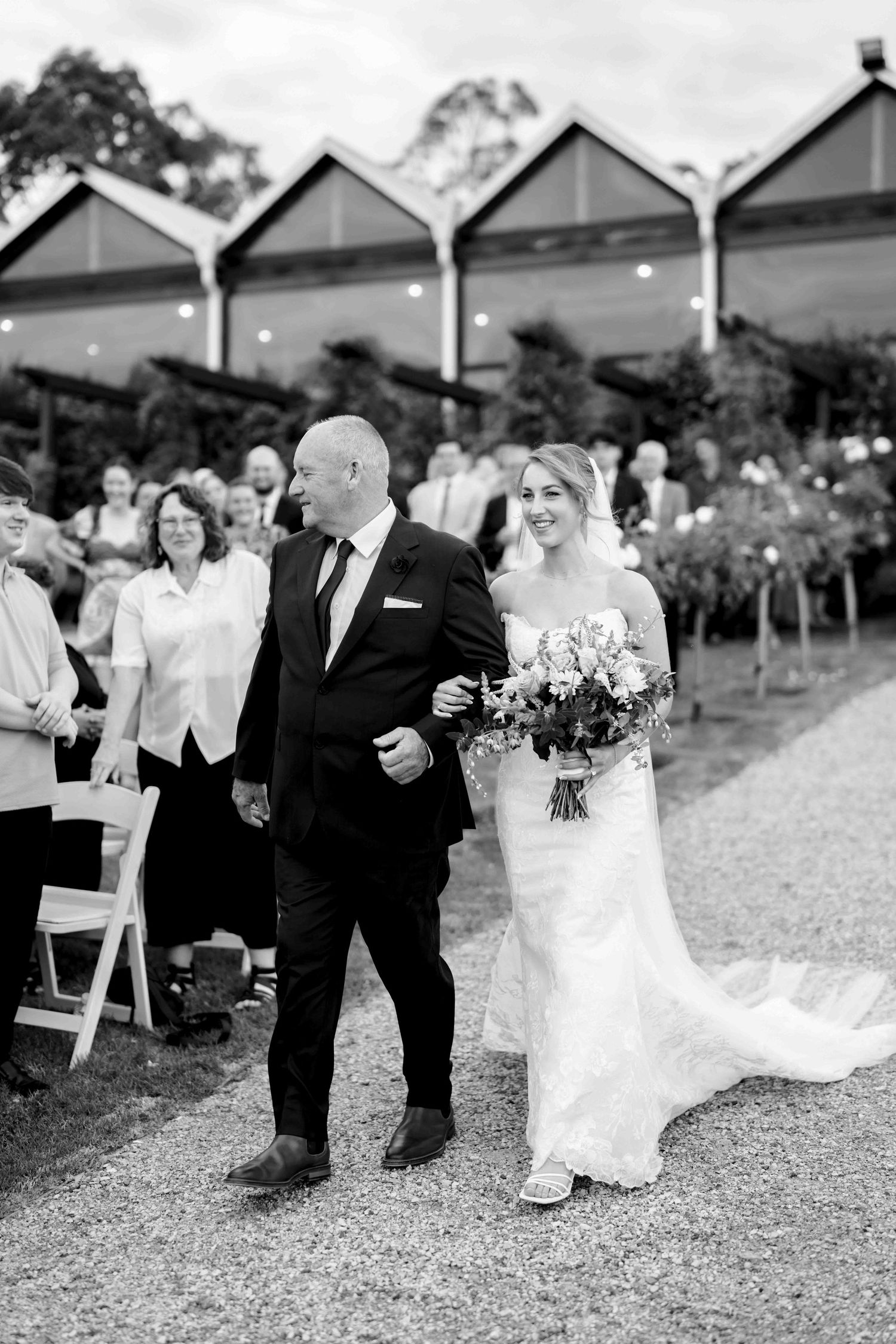 Black and white photo of bride walking down the aisle with her father at an outdoor wedding venue.