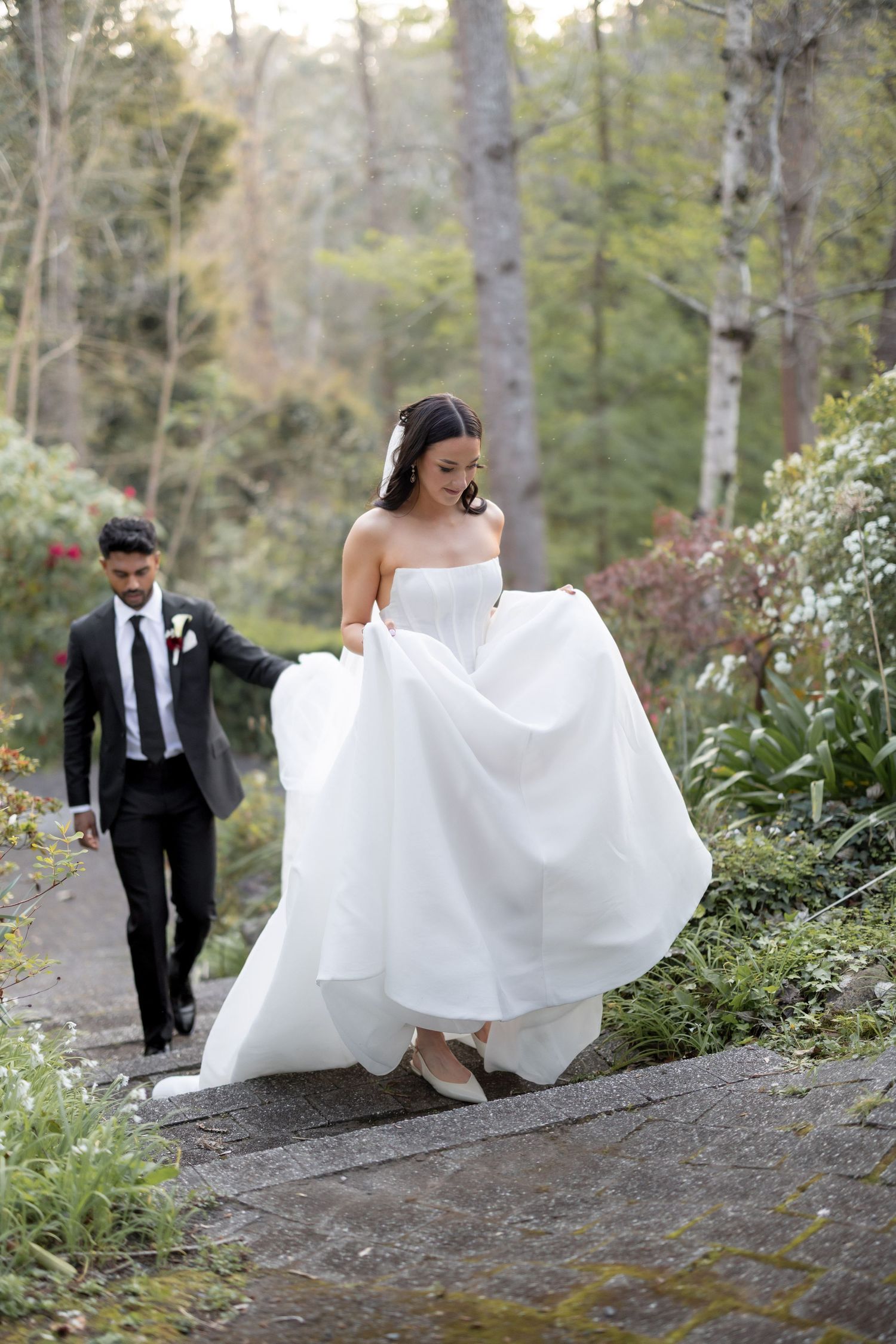 Bride in white strapless gown lifts dress walking stone path outdoors, groom in black suit follows through lush garden.