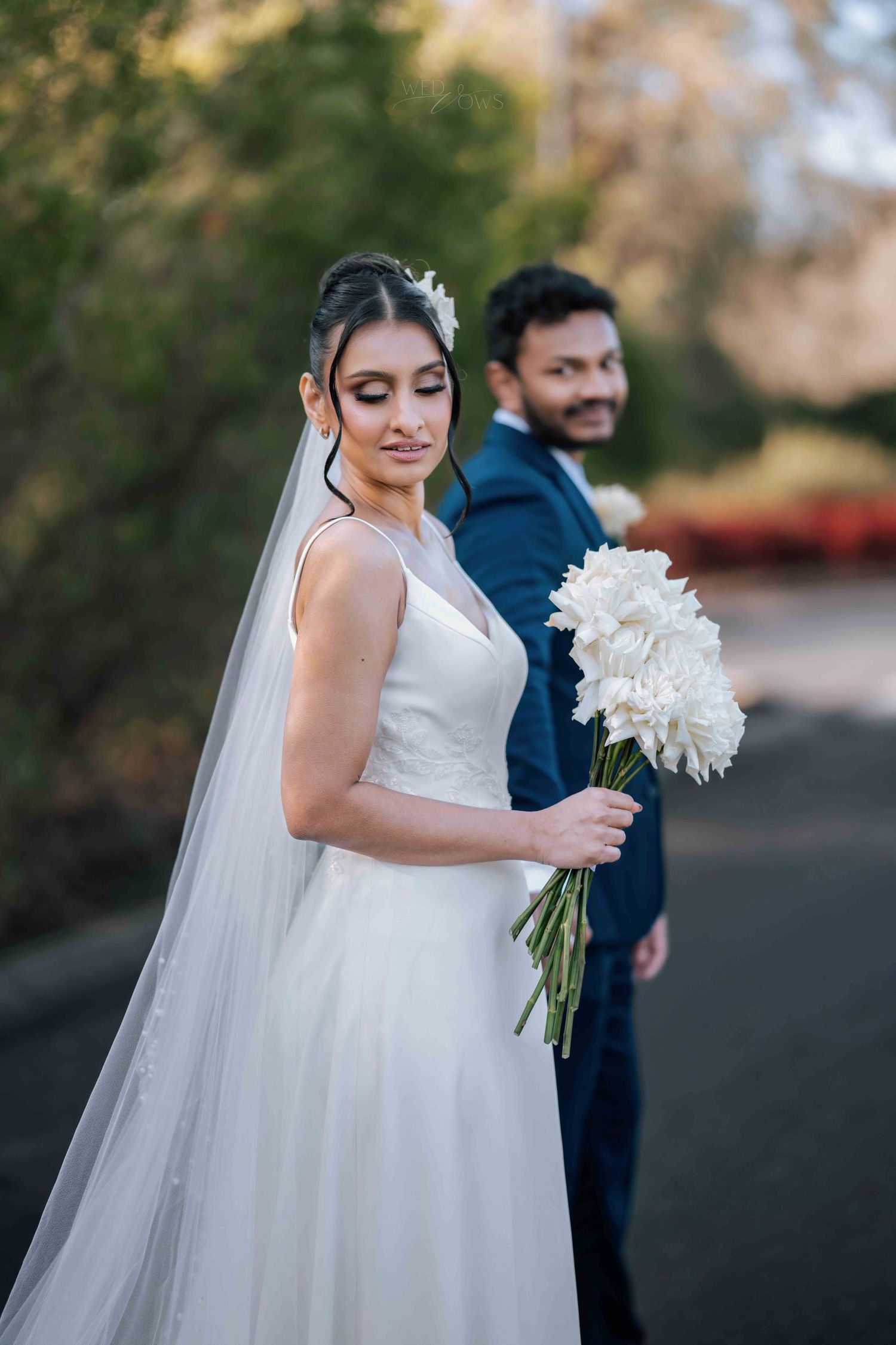 Bride in white ballgown holding white peony bouquet with groom in navy suit in background outdoors.