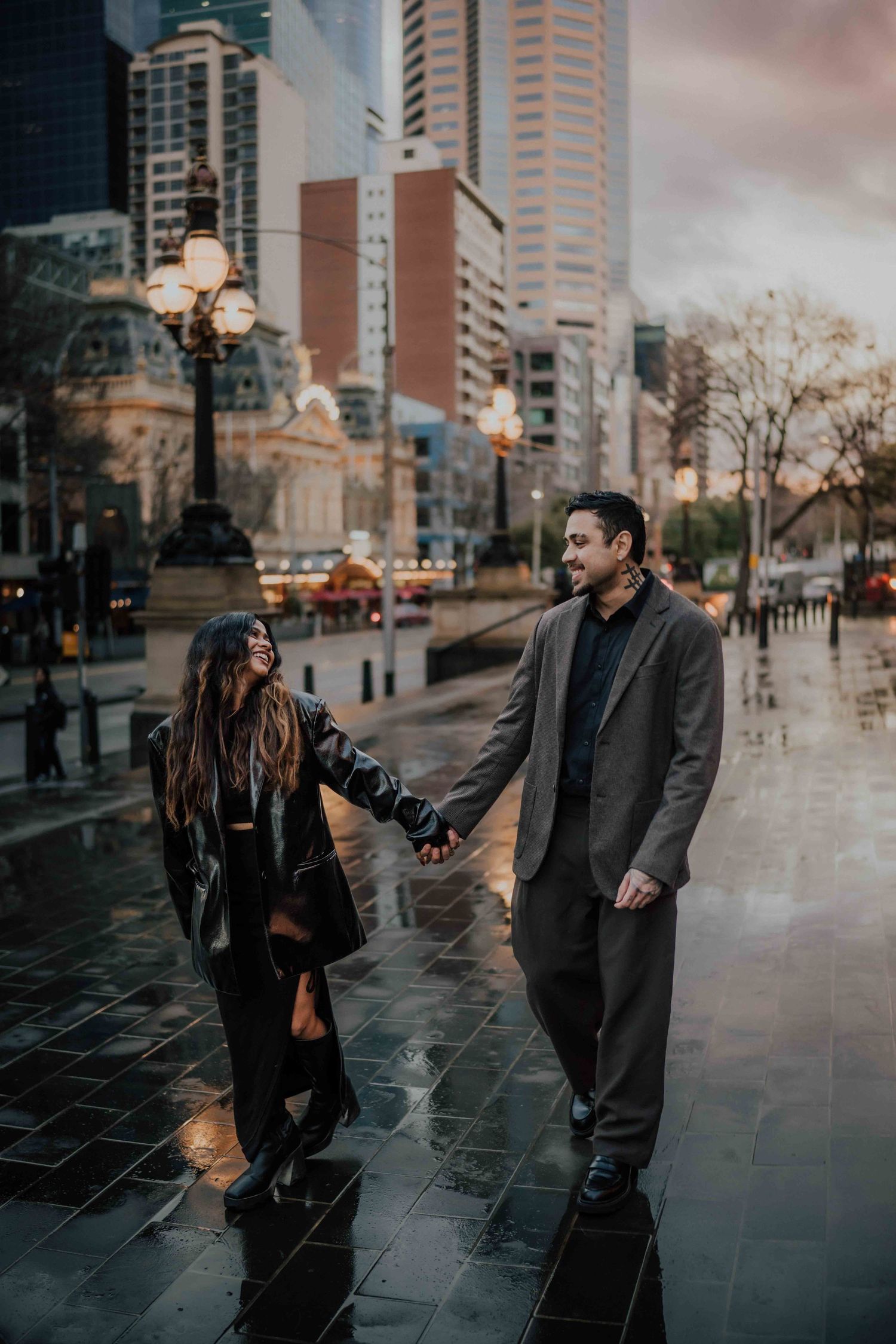 Happy couple holding hands and laughing while walking on a wet city street at dusk.