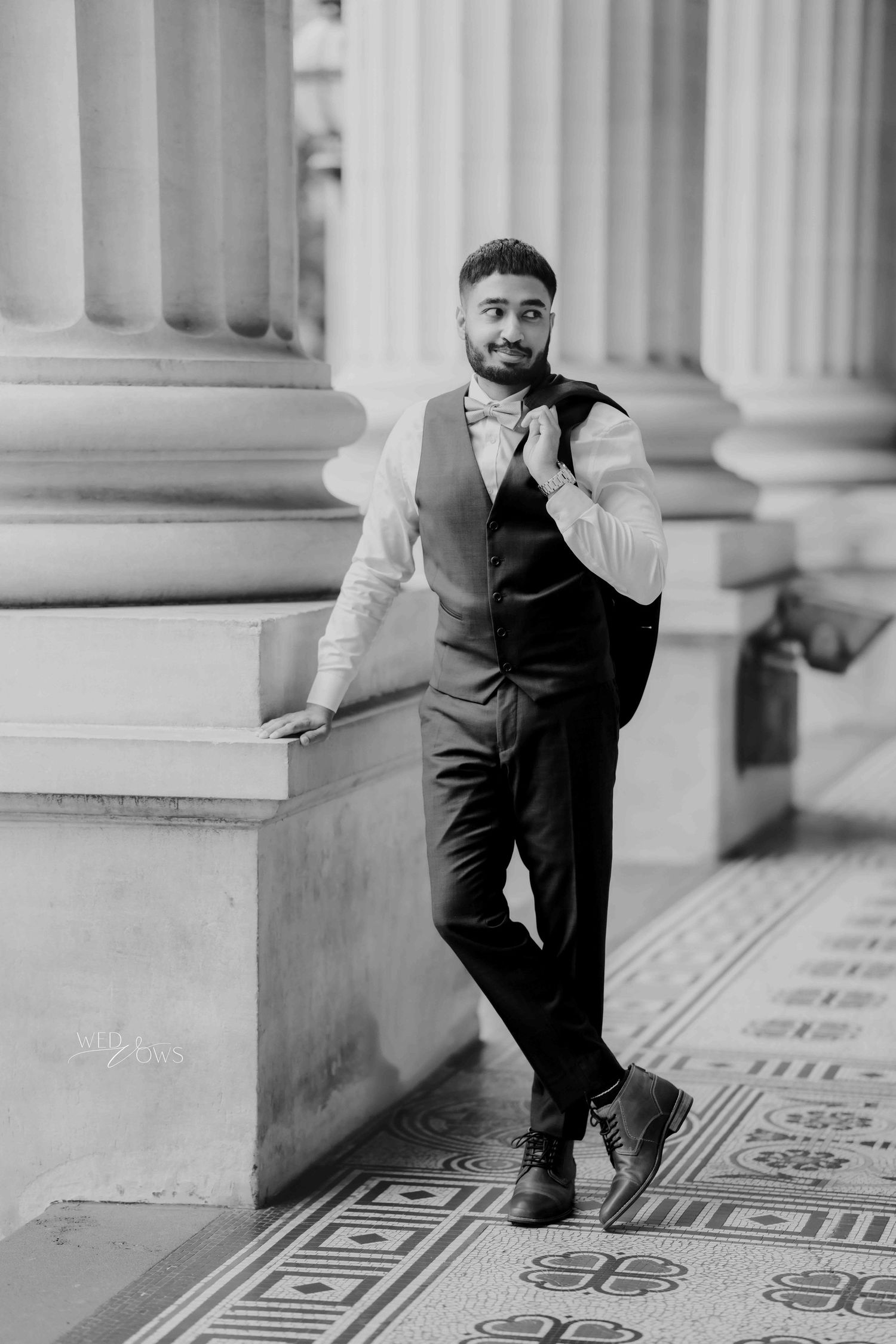 Stylish groom in black vest and bow tie leans against stone column in black and white photo.