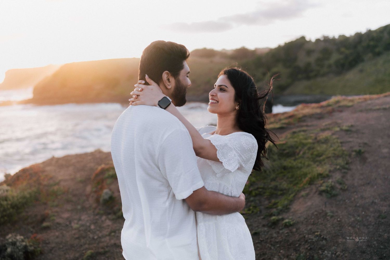Couple in white outfits holding each other by the ocean at sunset on rocky coastline.