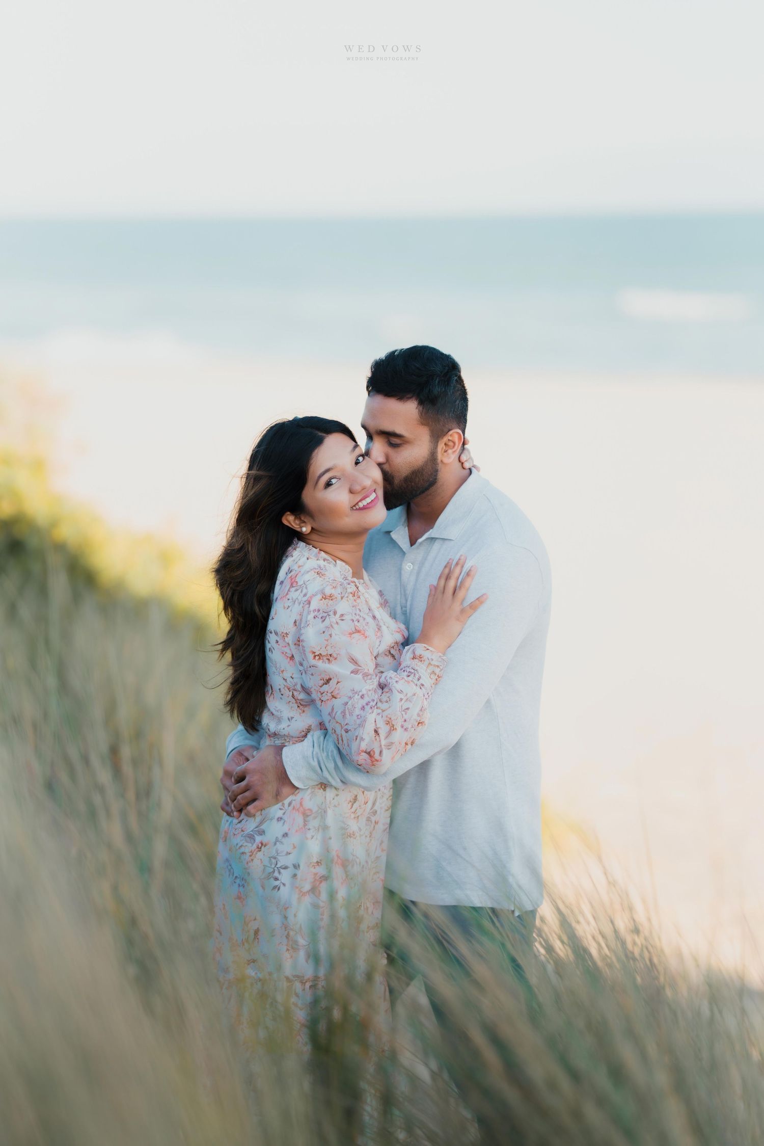 Couple embracing among tall coastal grass with ocean in background during soft golden hour light.