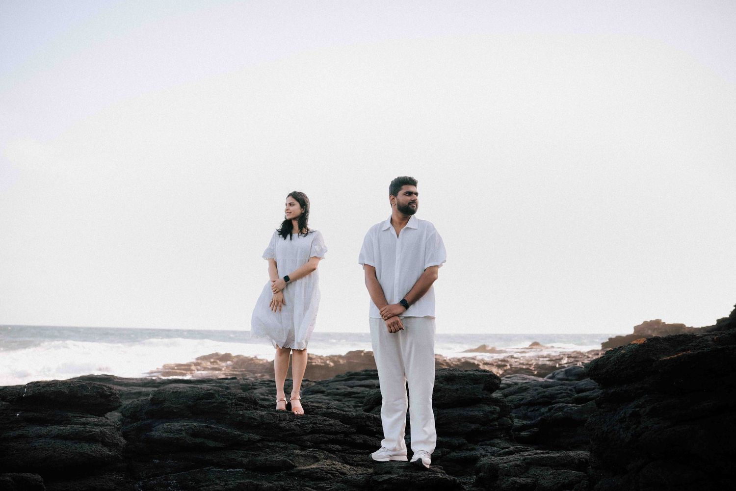 Couple standing apart on coastal rocks in white outfits with misty ocean behind them.