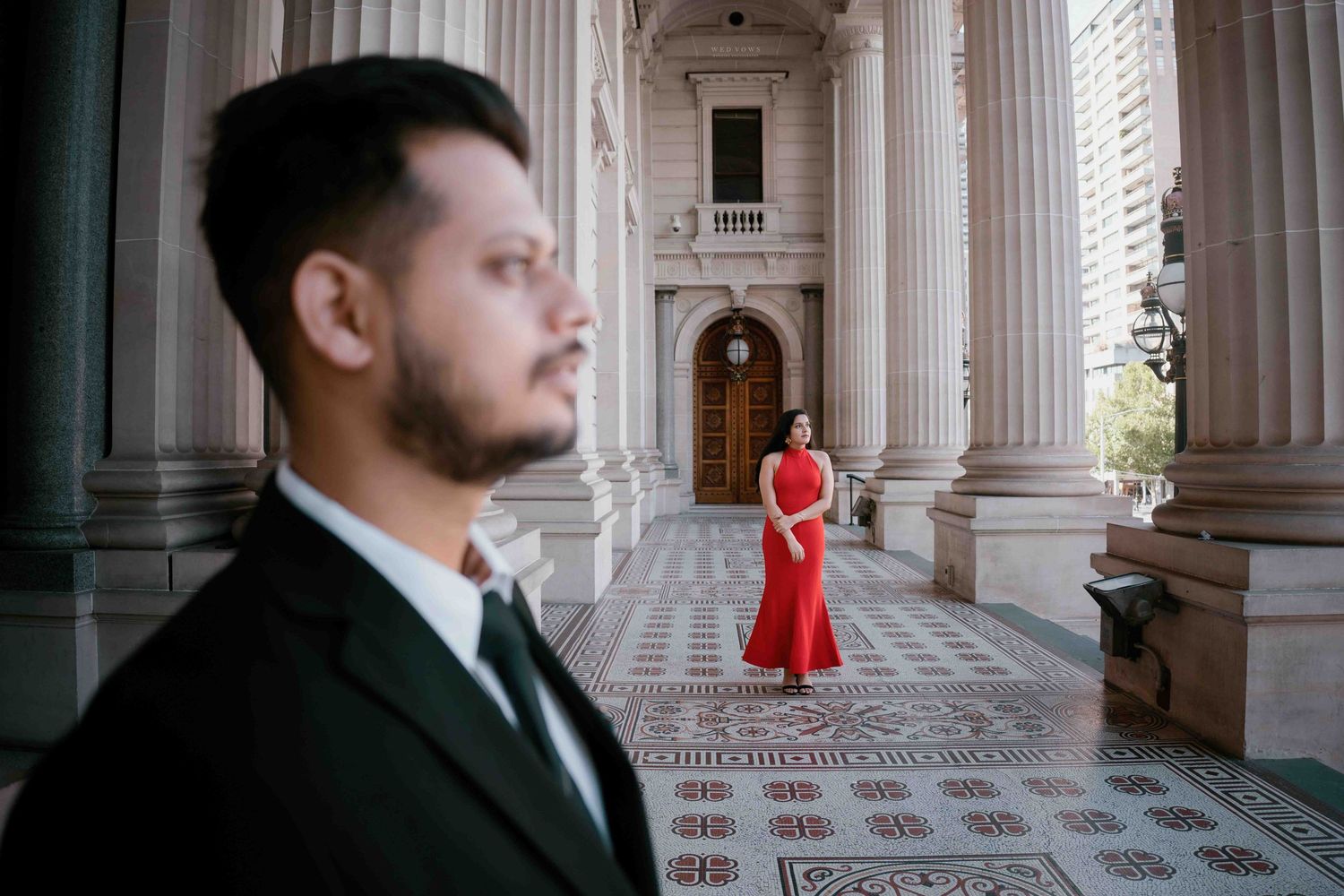Woman in red gown poses between classical columns as man in dark suit stands in foreground.
