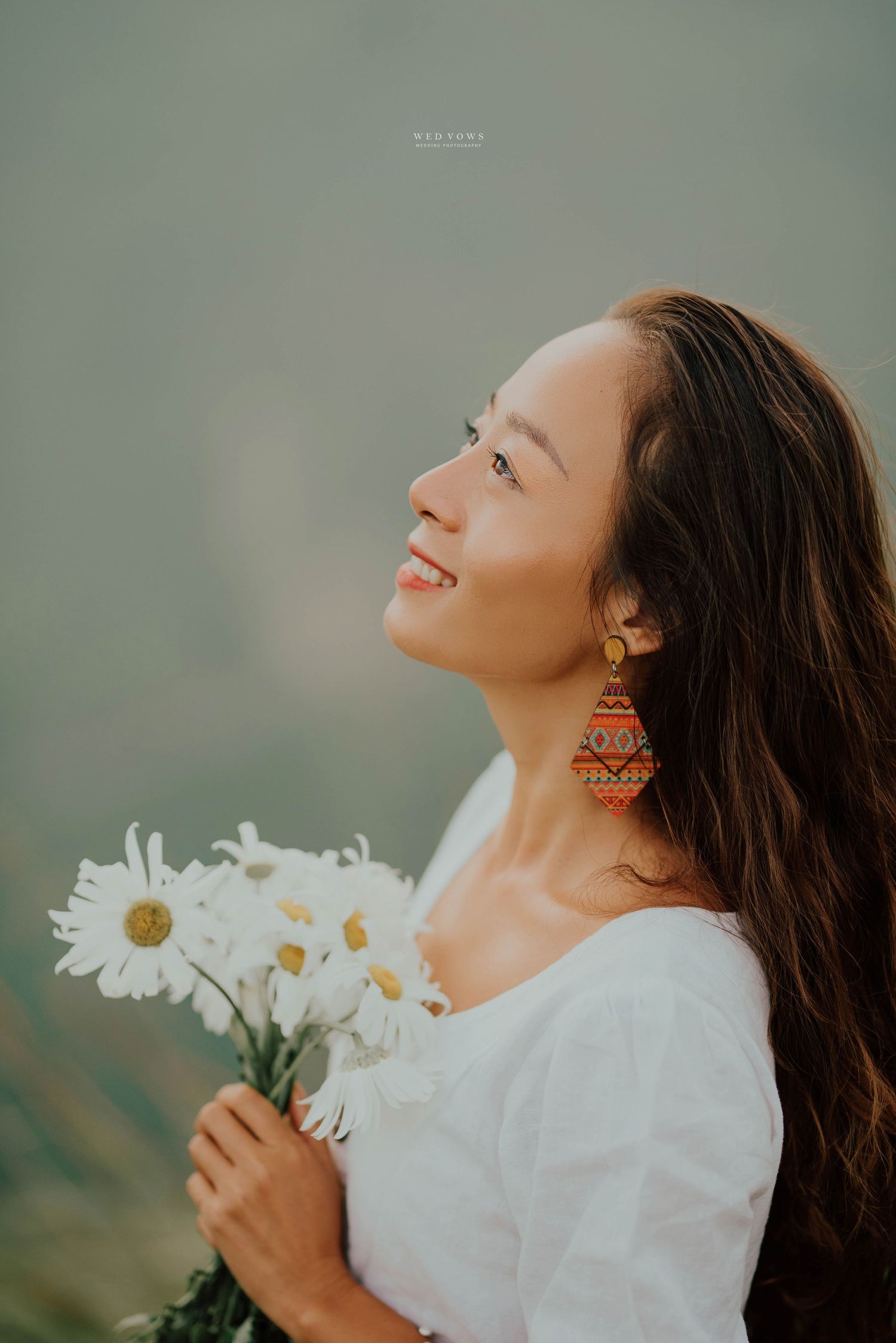 A smiling young woman with long hair holds white daisy flowers, wearing colorful earrings and white top.