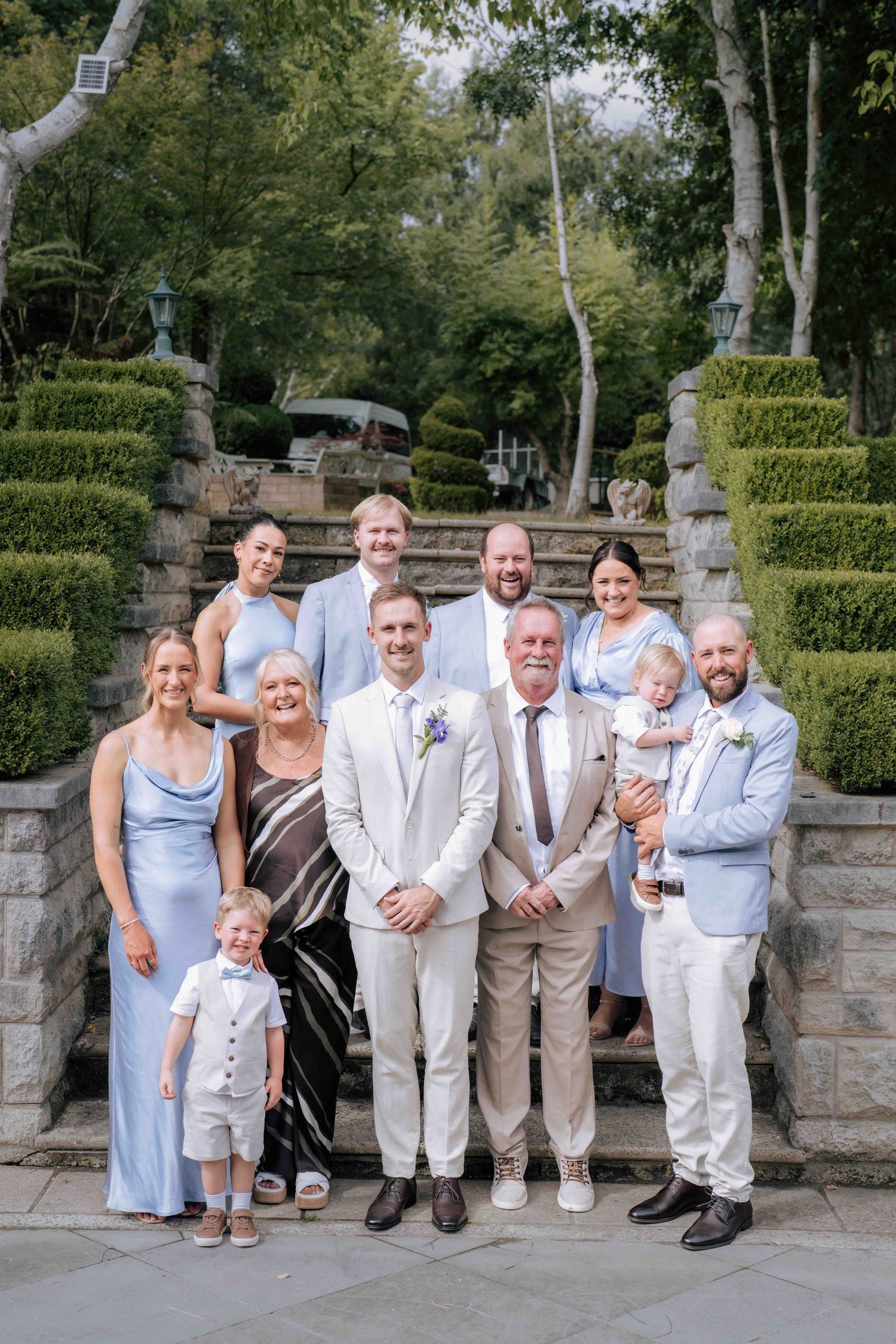 Groom poses with extended family group on stone staircase at elegant garden wedding.