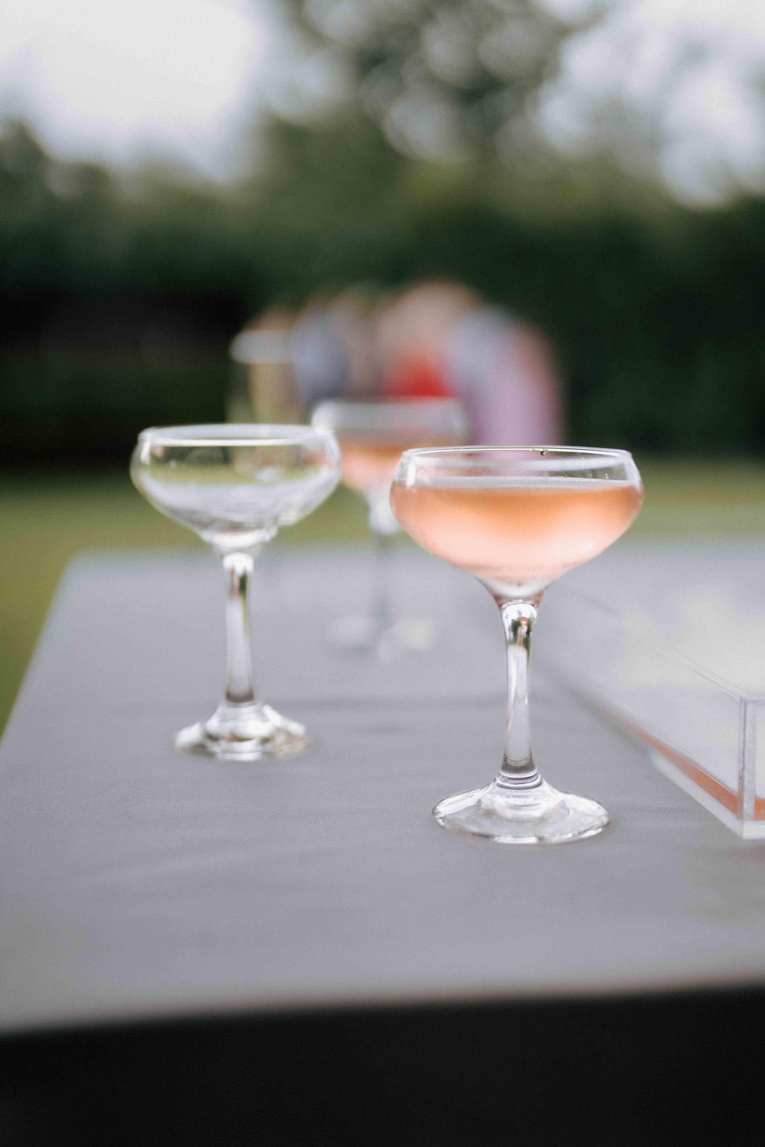 Two elegant coupe glasses with rose champagne on a gray linen table at an outdoor wedding reception.