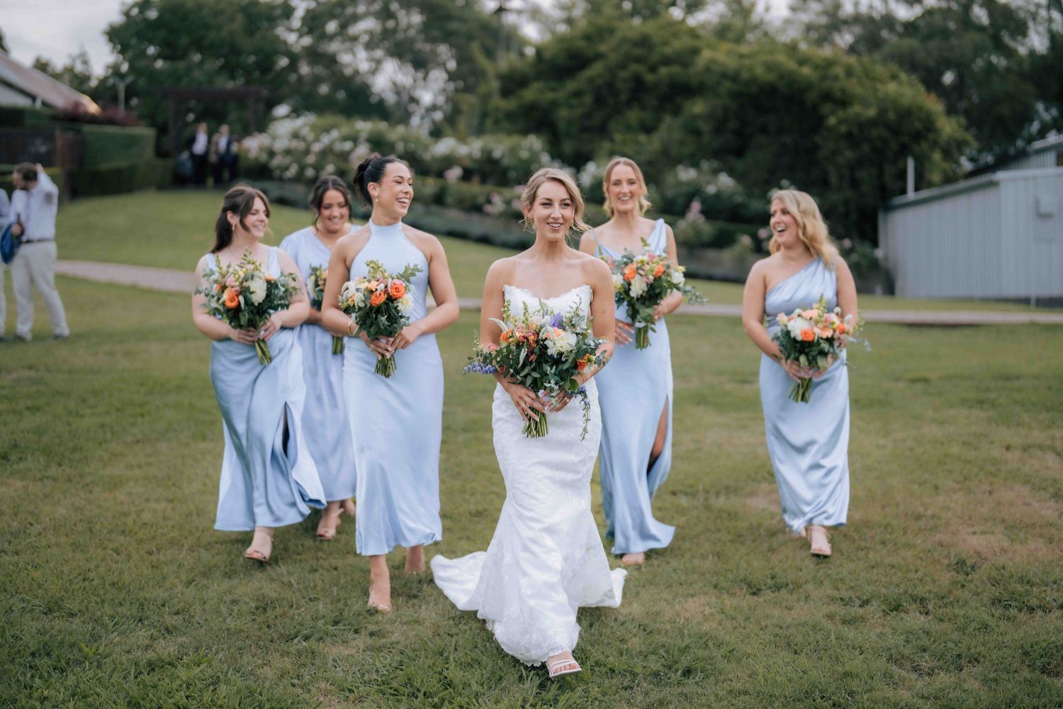 Bride leads bridesmaids in powder blue dresses across green lawn at outdoor wedding.