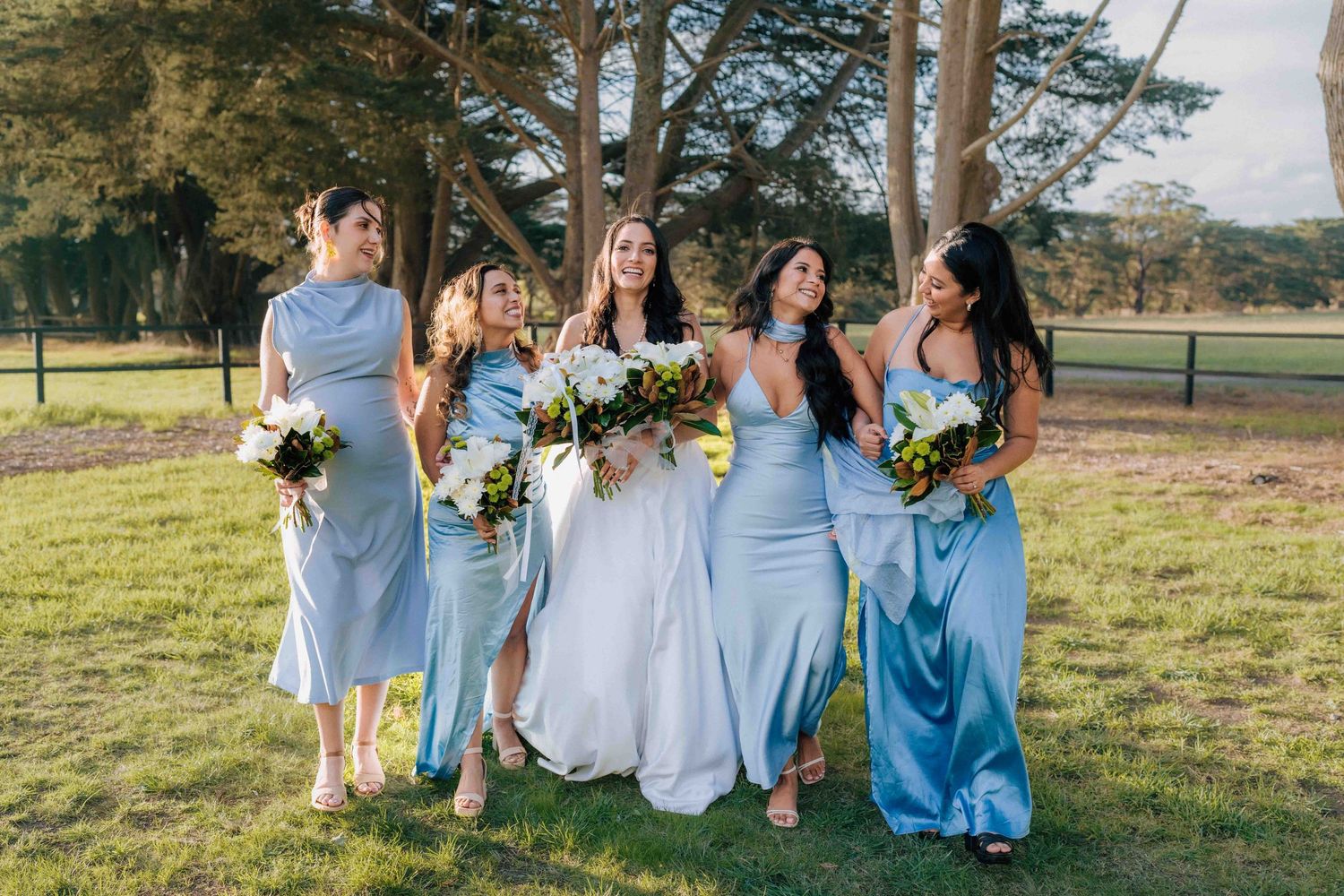 Bride with four bridesmaids in light blue gowns holding bouquets outdoors on wedding day.