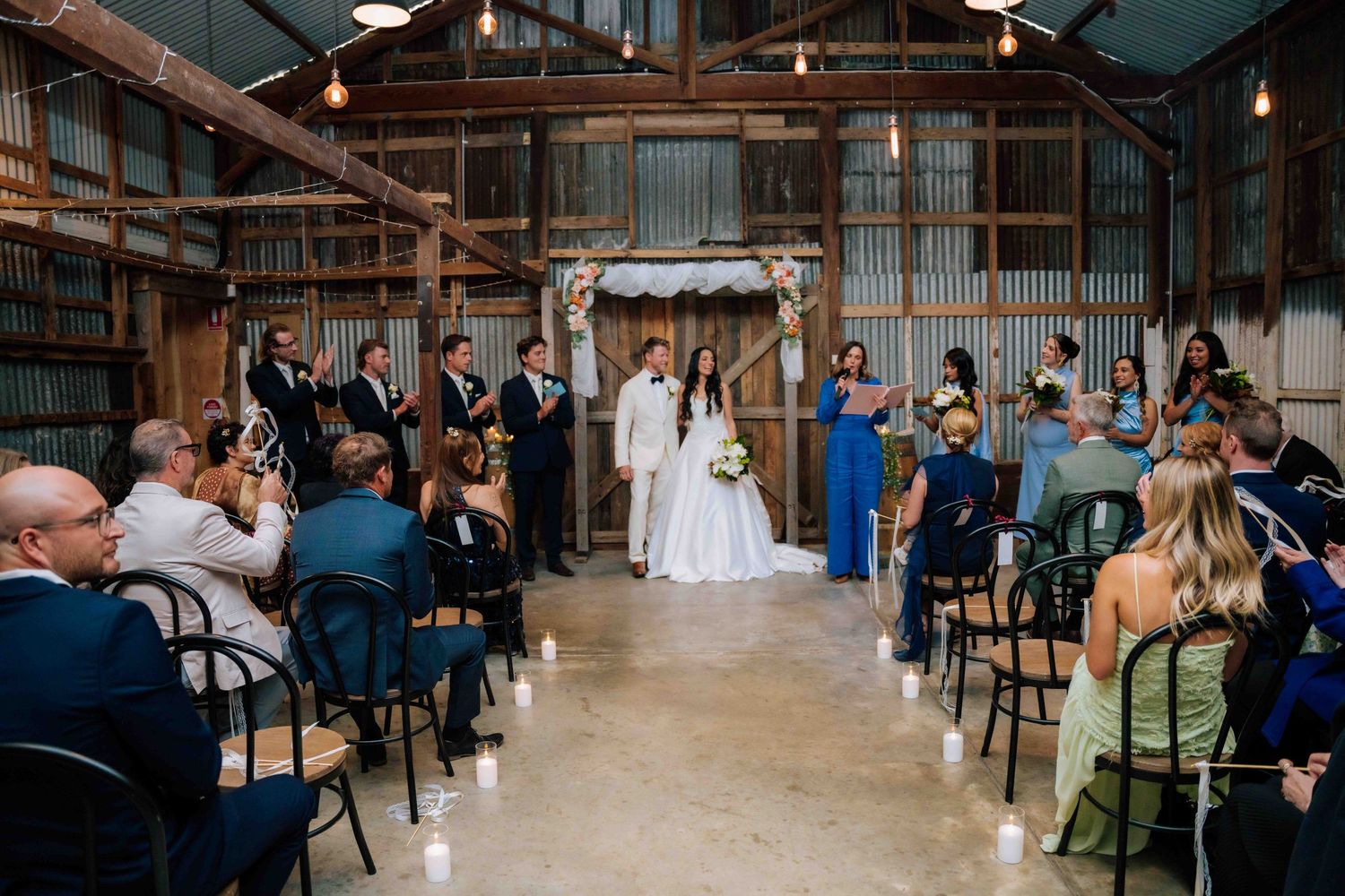 Bride and groom exchange vows at rustic barn wedding ceremony with floral arch, guests seated in black chairs.