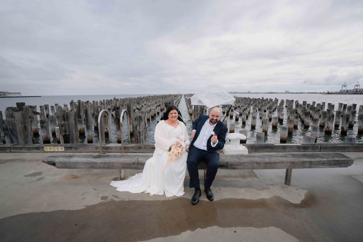 Bride and groom posing playfully on a rainy pier with weathered wooden pylons in the background.