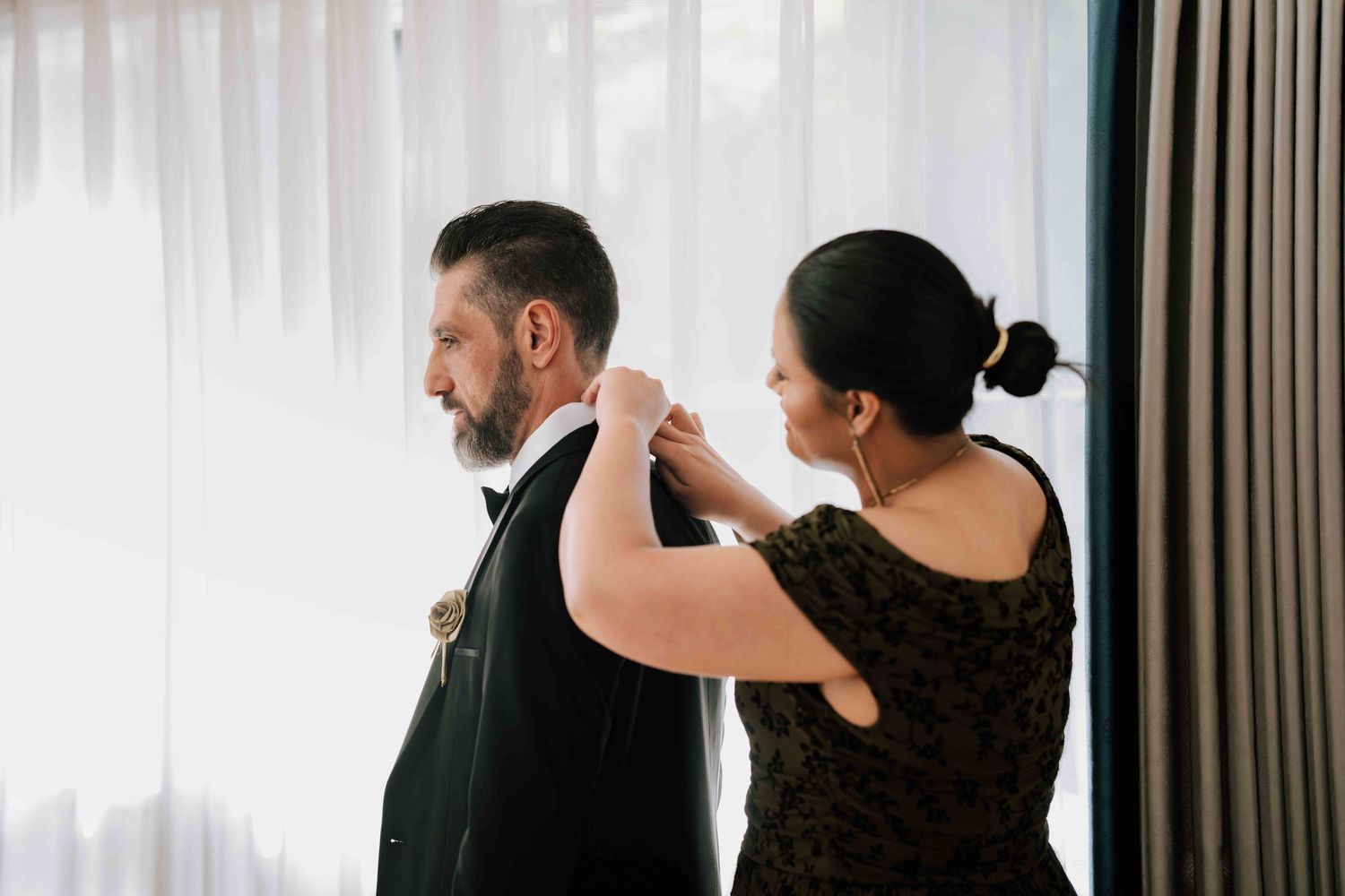 Woman in dark floral dress adjusts groom's tuxedo collar from behind near sheer curtained window.