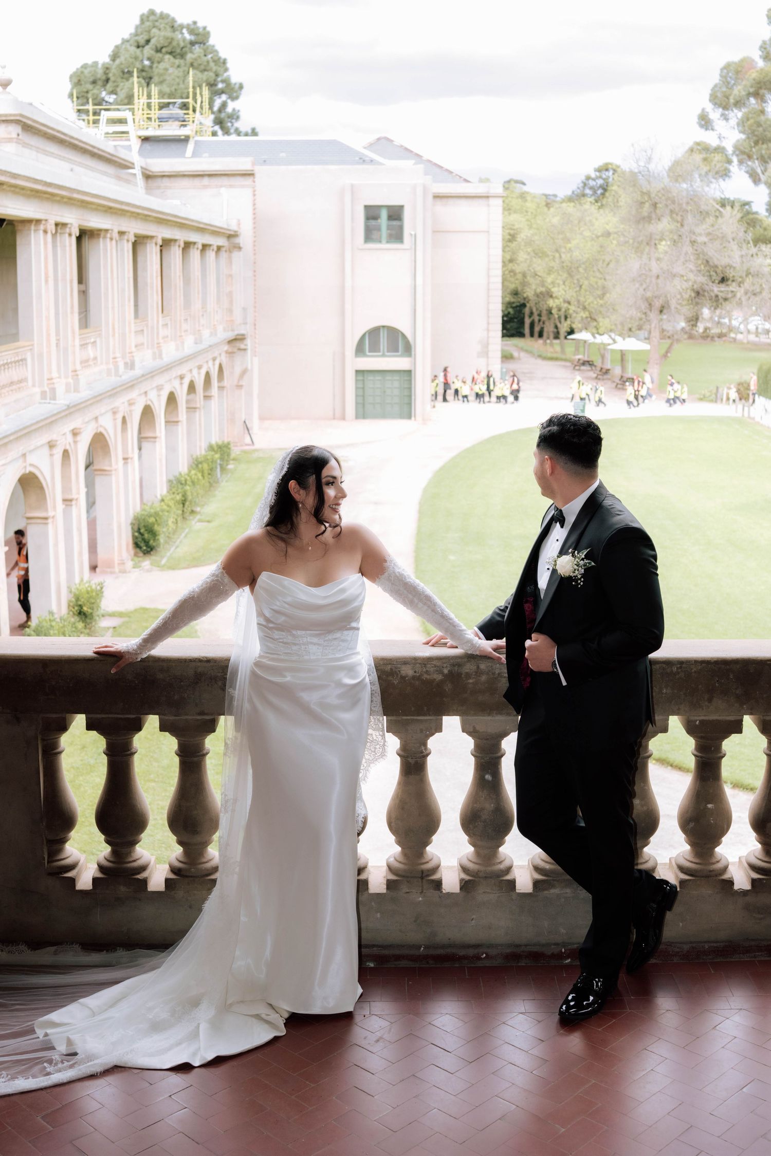 Bride and groom share a romantic moment on a grand balcony overlooking lush wedding venue grounds.