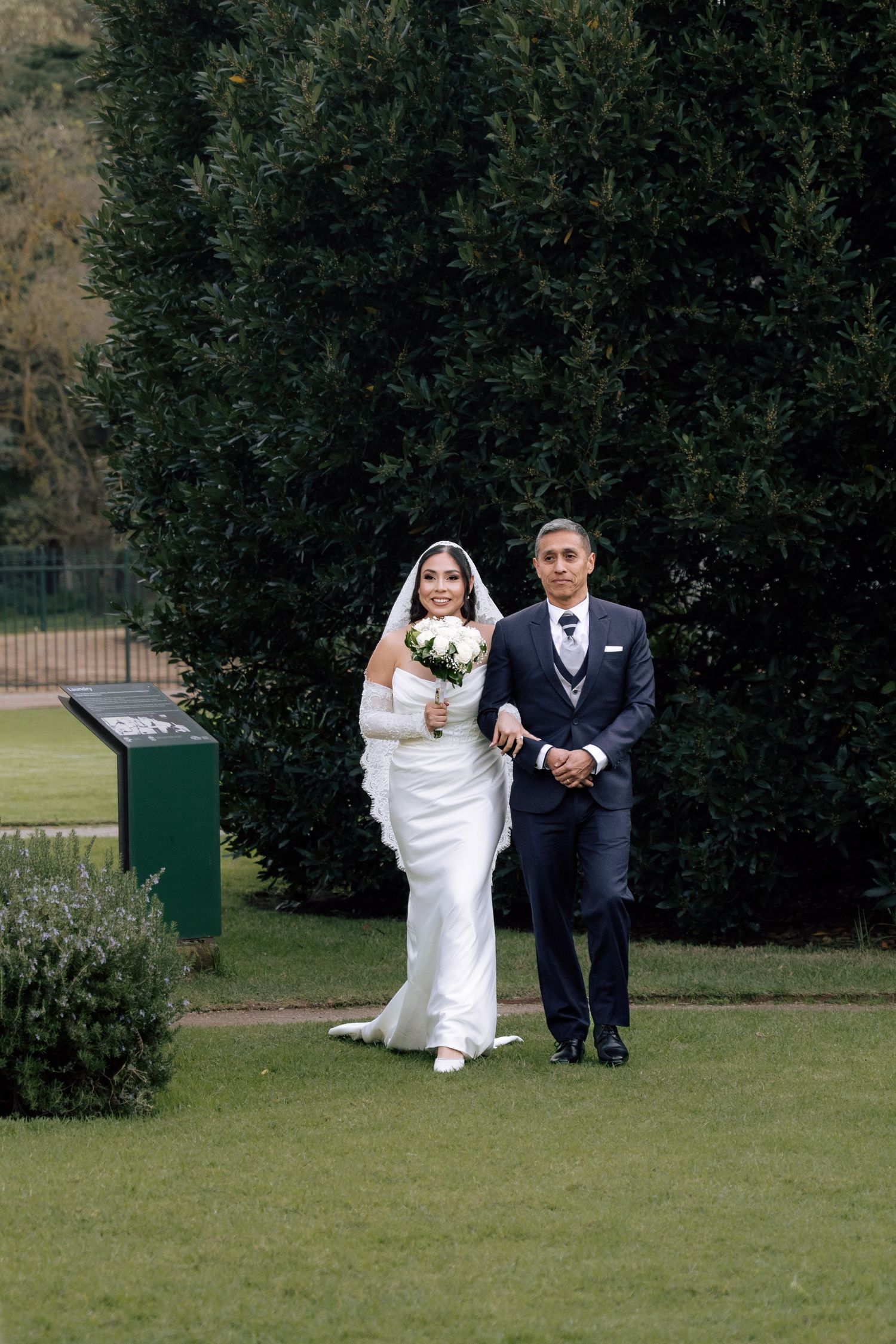 Bride in white gown with veil holding bouquet walking with escort through garden.