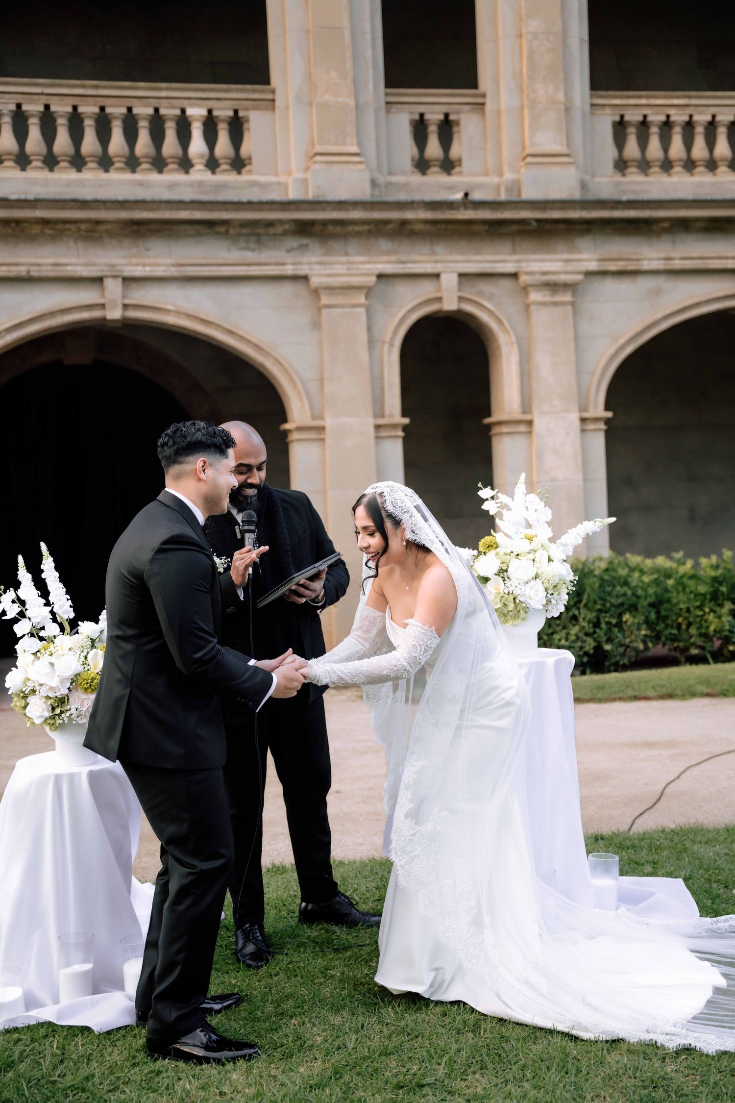 Bride and groom exchange vows outdoors at elegant wedding ceremony with white floral arrangements and stone arched building.
