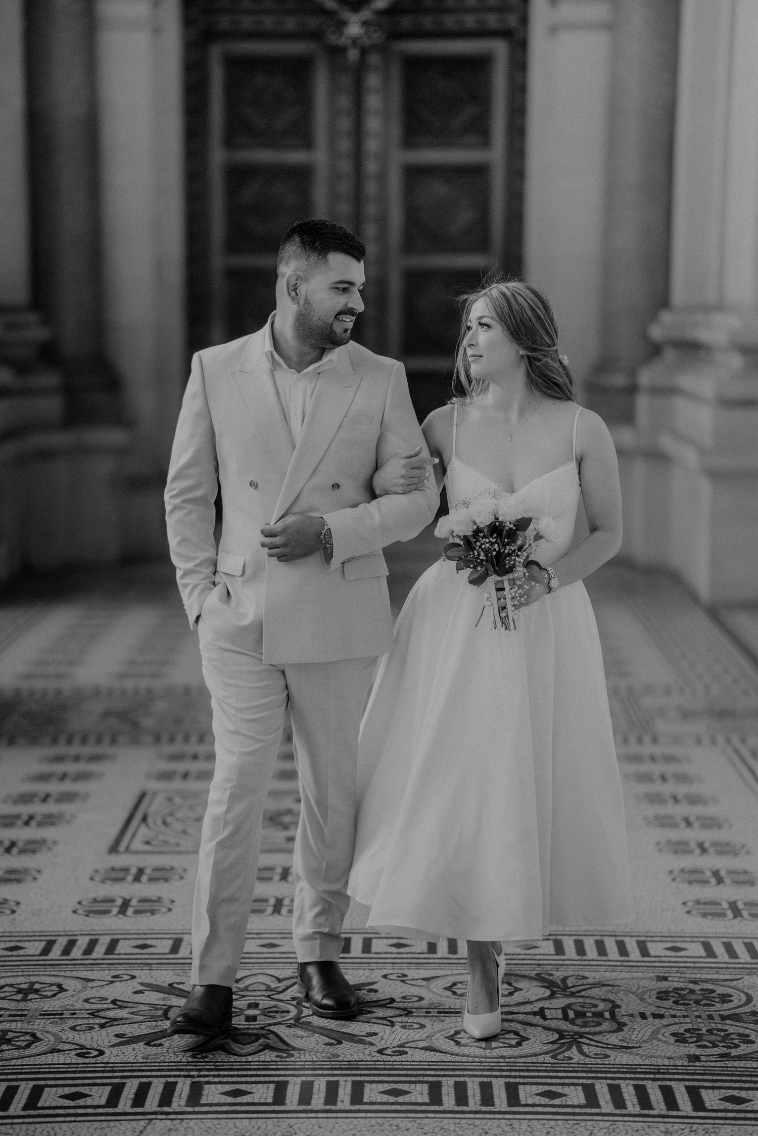 Black and white photo of a smiling bride and groom walking together in an elegant architectural setting.