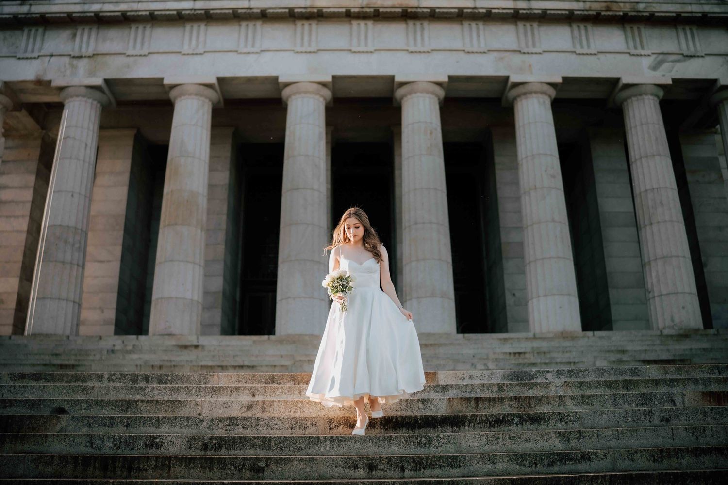 Bride in white midi gown with bouquet standing on stone steps before grand columned building.