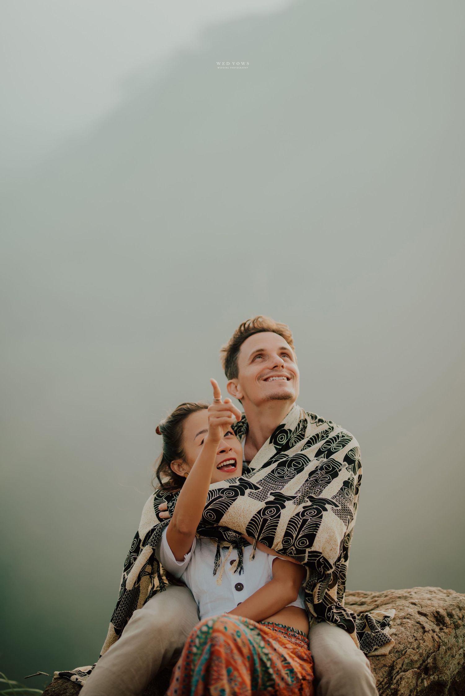 Happy couple sitting on rocks in misty mountains, woman pointing upward joyfully.