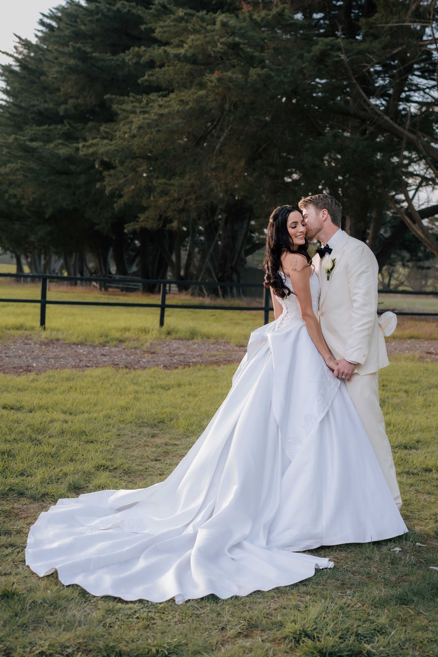 Groom in ivory suit kisses bride wearing a flowing white ball gown with long train in a garden setting.