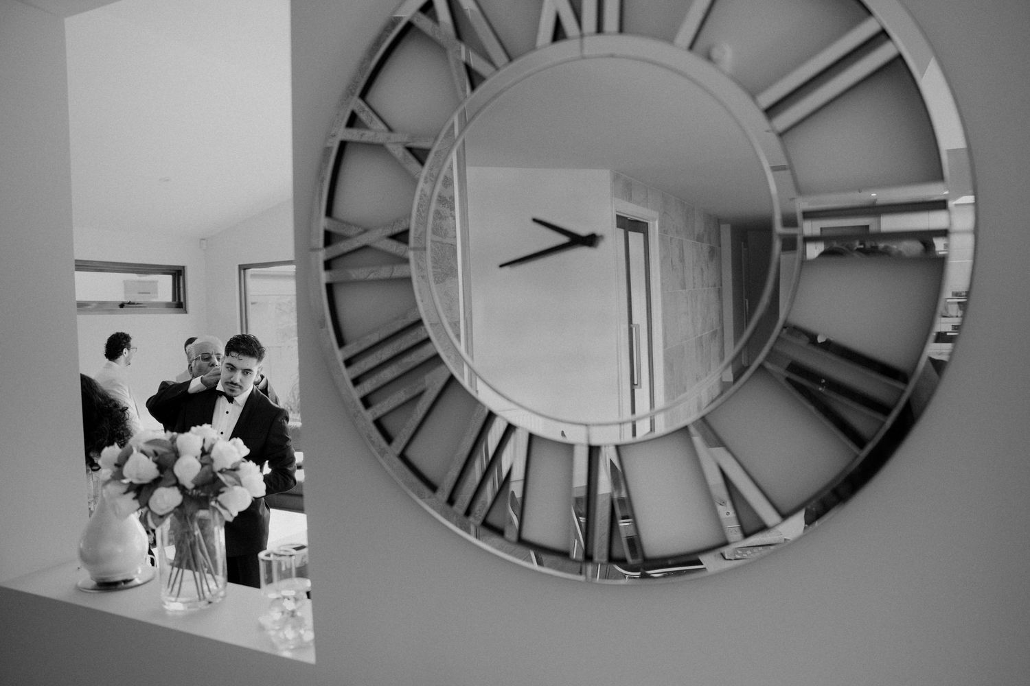 Groom preparations reflected in large decorative round mirror with Roman numerals in black and white.