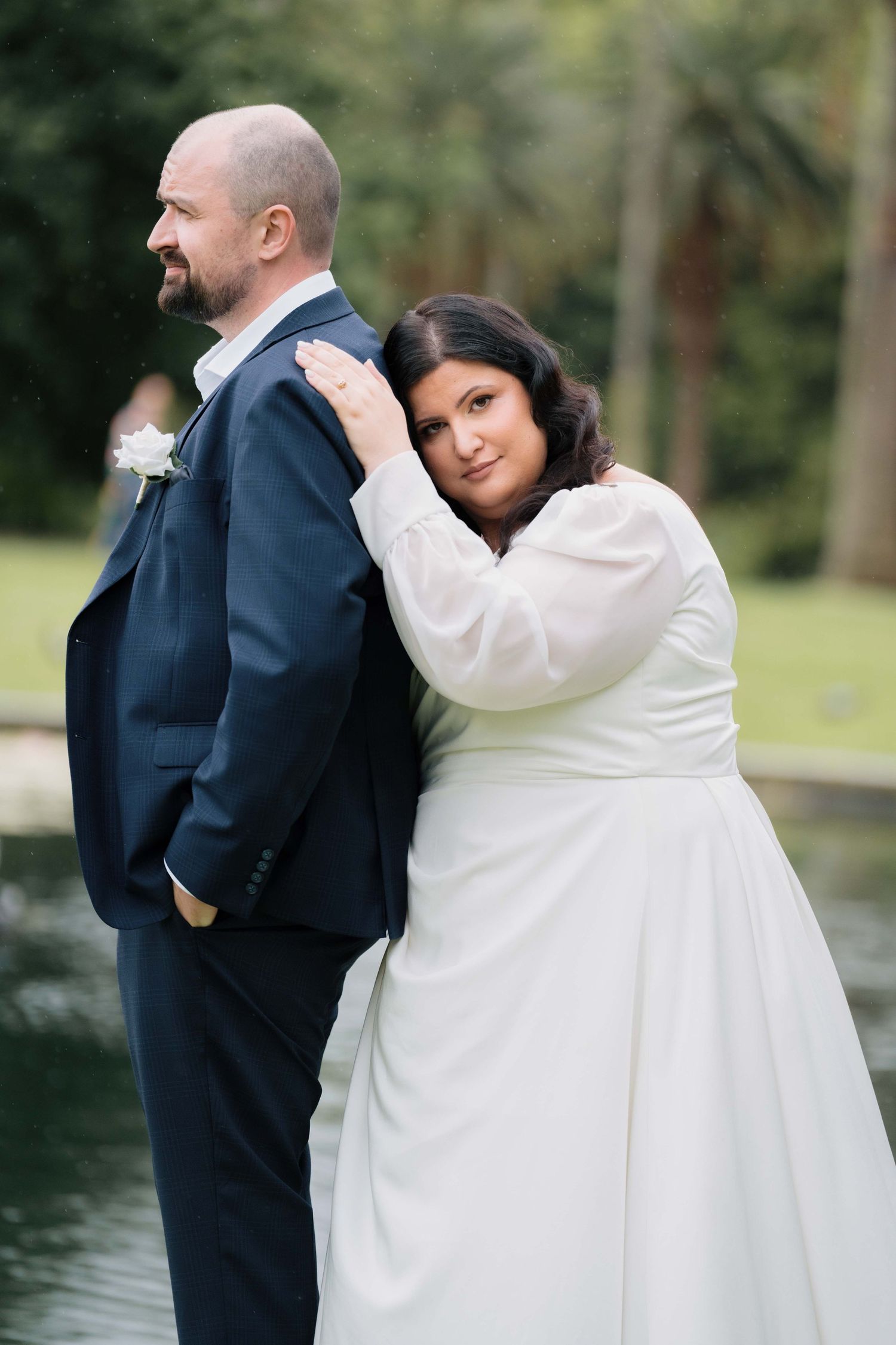 Bride leans on grooms shoulder from behind near a pond, wearing elegant white long-sleeve wedding gown.