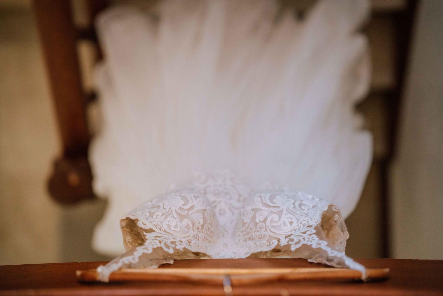 Delicate lace bridal garter displayed on wooden chair with white tulle wedding dress behind.