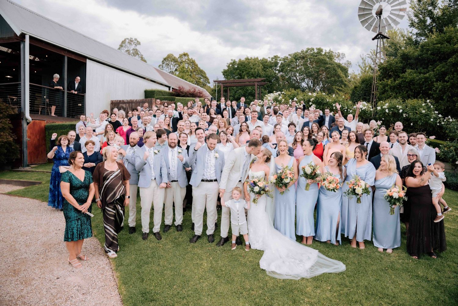 Joyful large wedding party group photo outside barn venue with windmill, guests cheering and celebrating together.